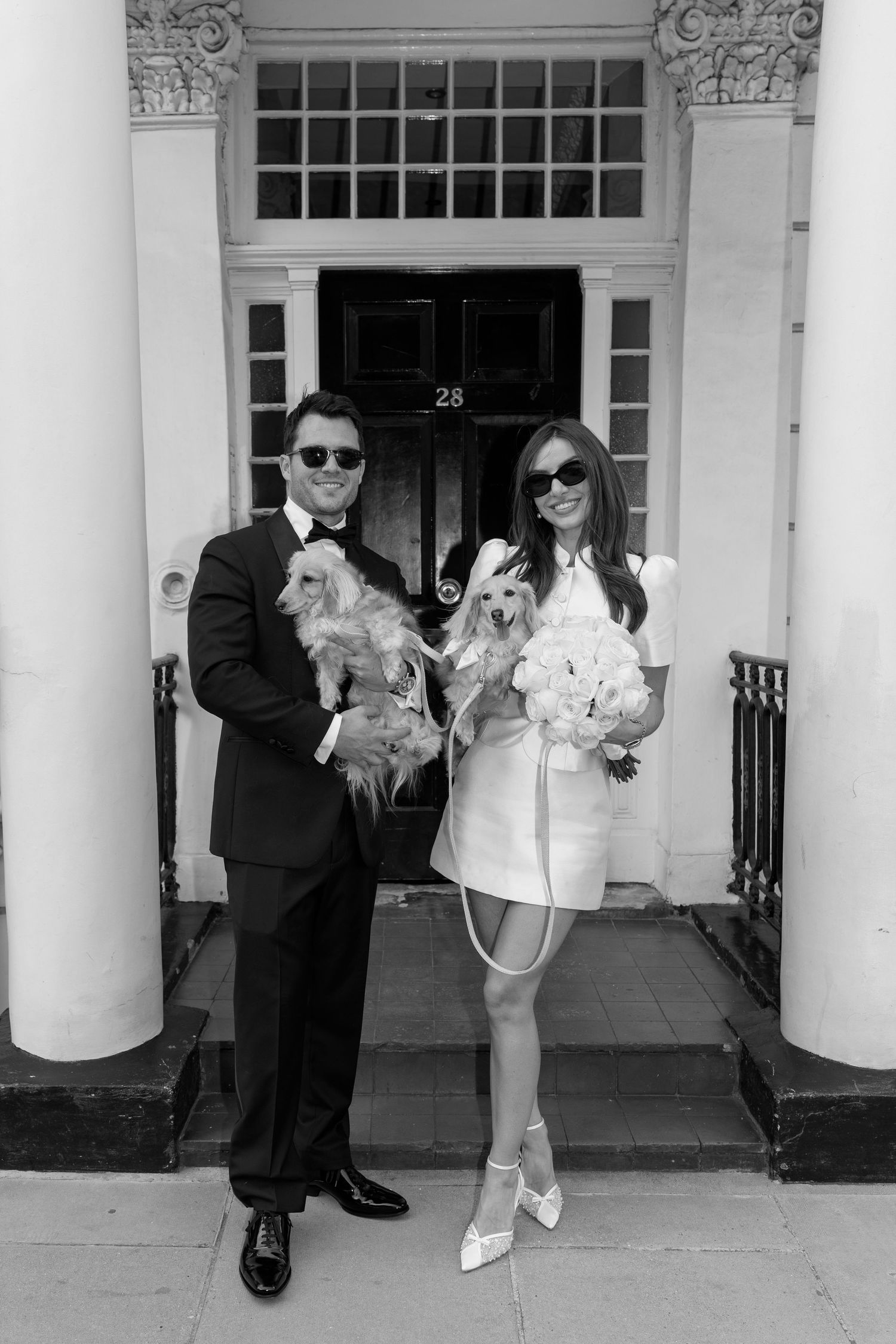 Black and white photo of bride and groom holding dogs outside a London townhouse door.