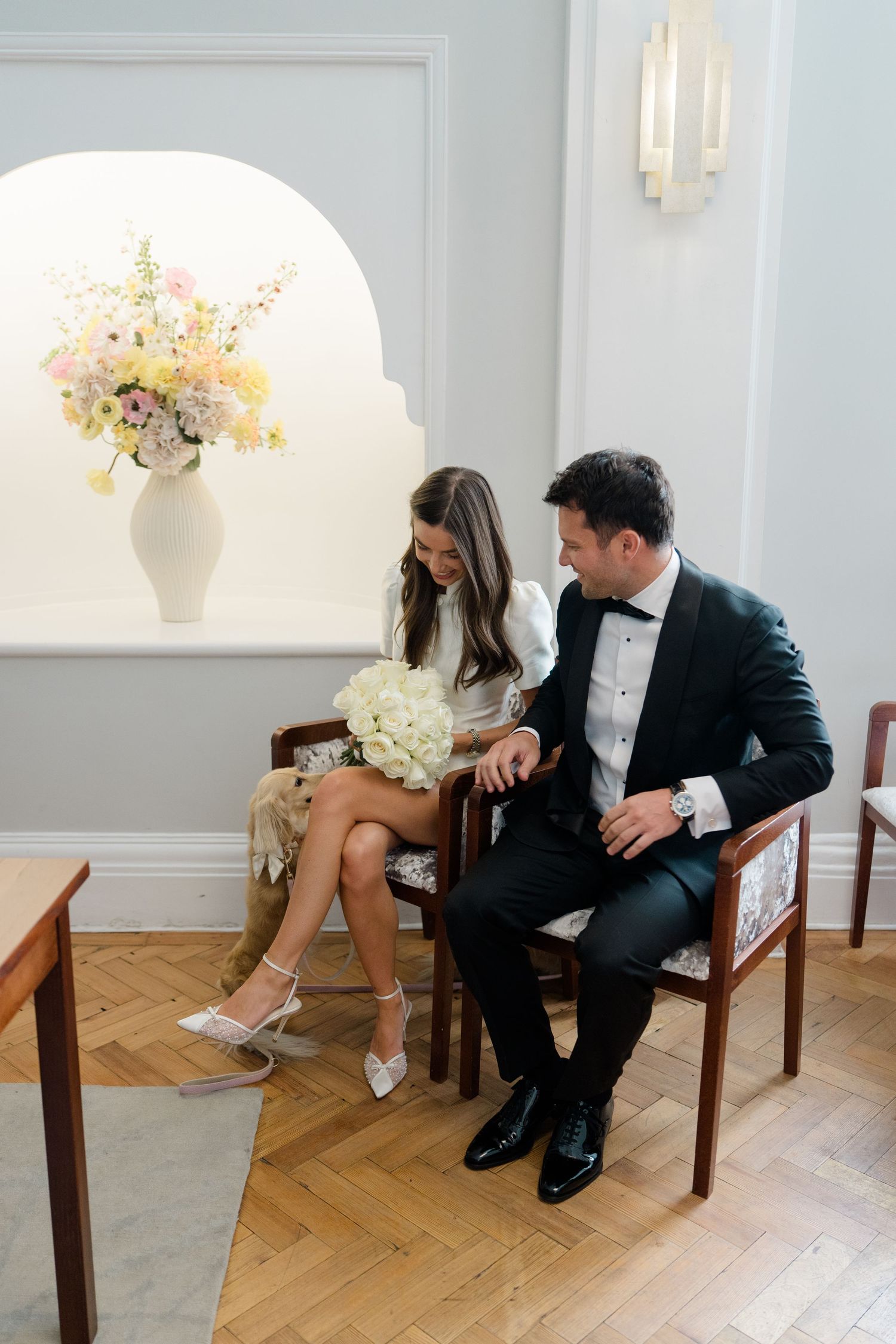 Bride and groom in elegant attire sitting together in a bright room with floral arrangement.