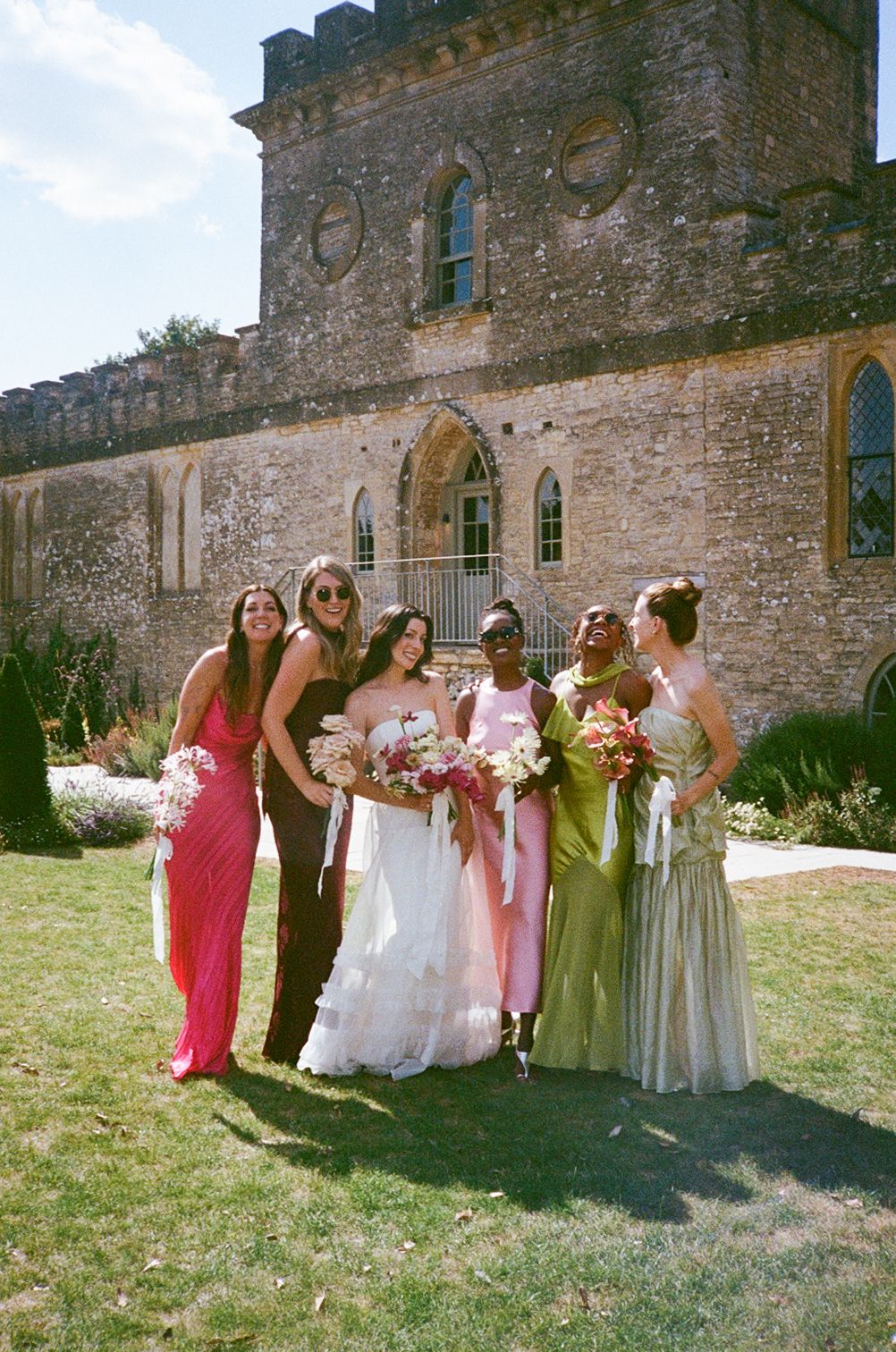 Bride in white gown poses with bridesmaids in colorful dresses outside a historic stone building on a sunny day.
