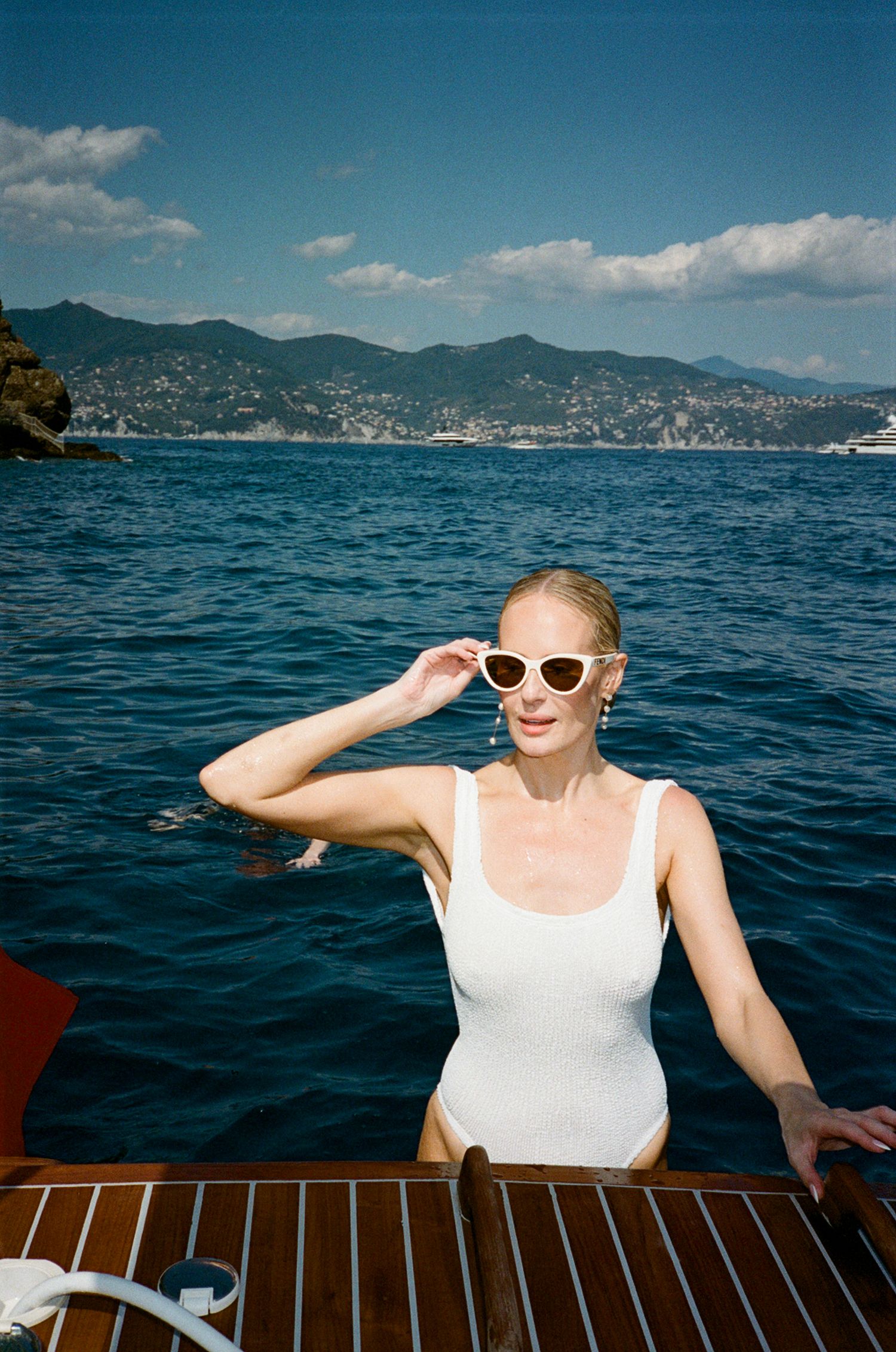 Woman in white swimsuit and sunglasses on a wooden boat with blue Mediterranean sea and mountains behind.