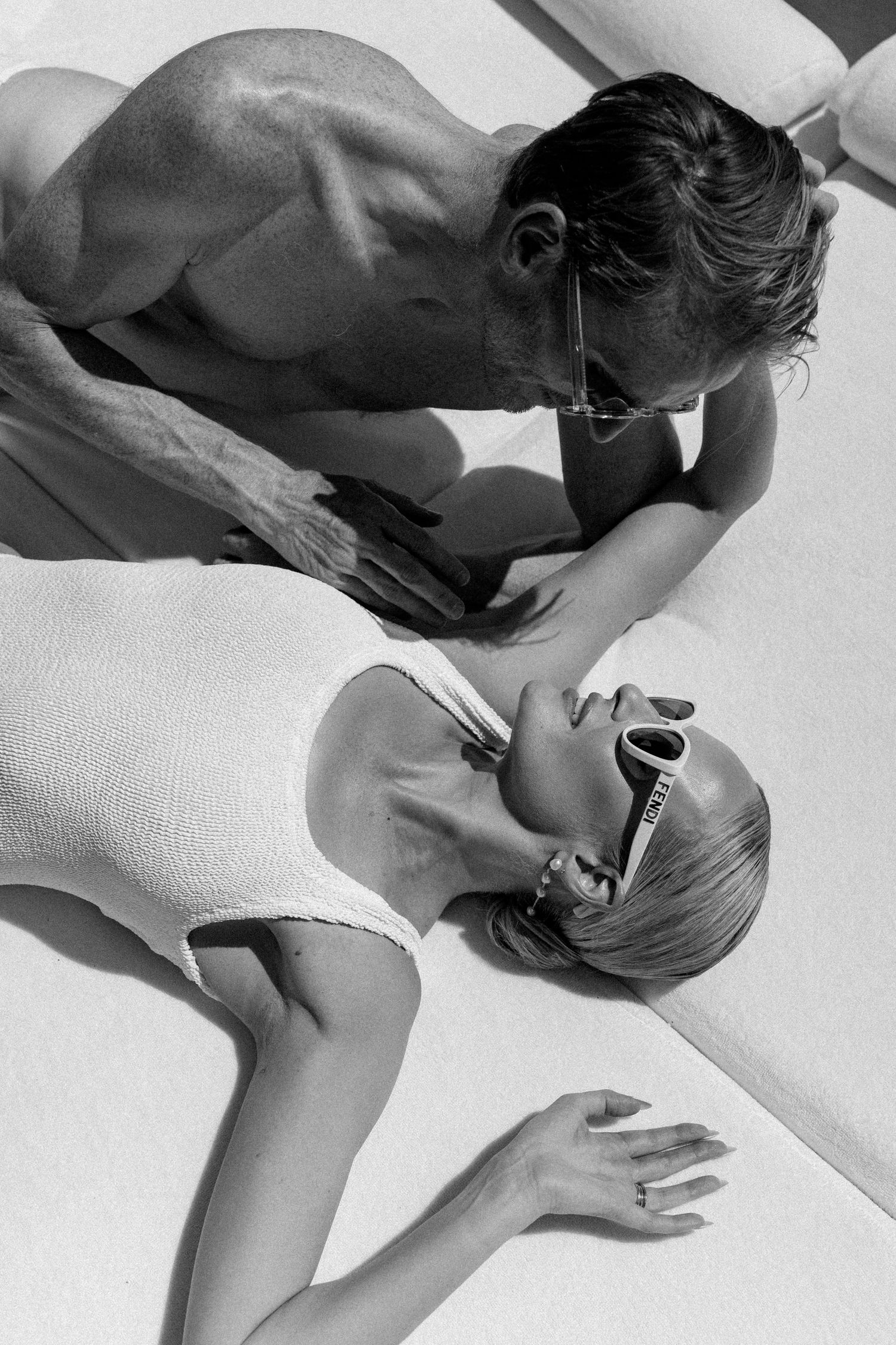 Black and white photo of a couple lounging poolside, woman in white swimsuit and sunglasses looking up at man.