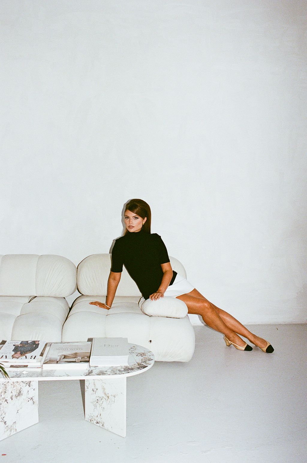 Woman in black top and white skirt poses on cream sofa beside marble coffee table in minimalist white room.