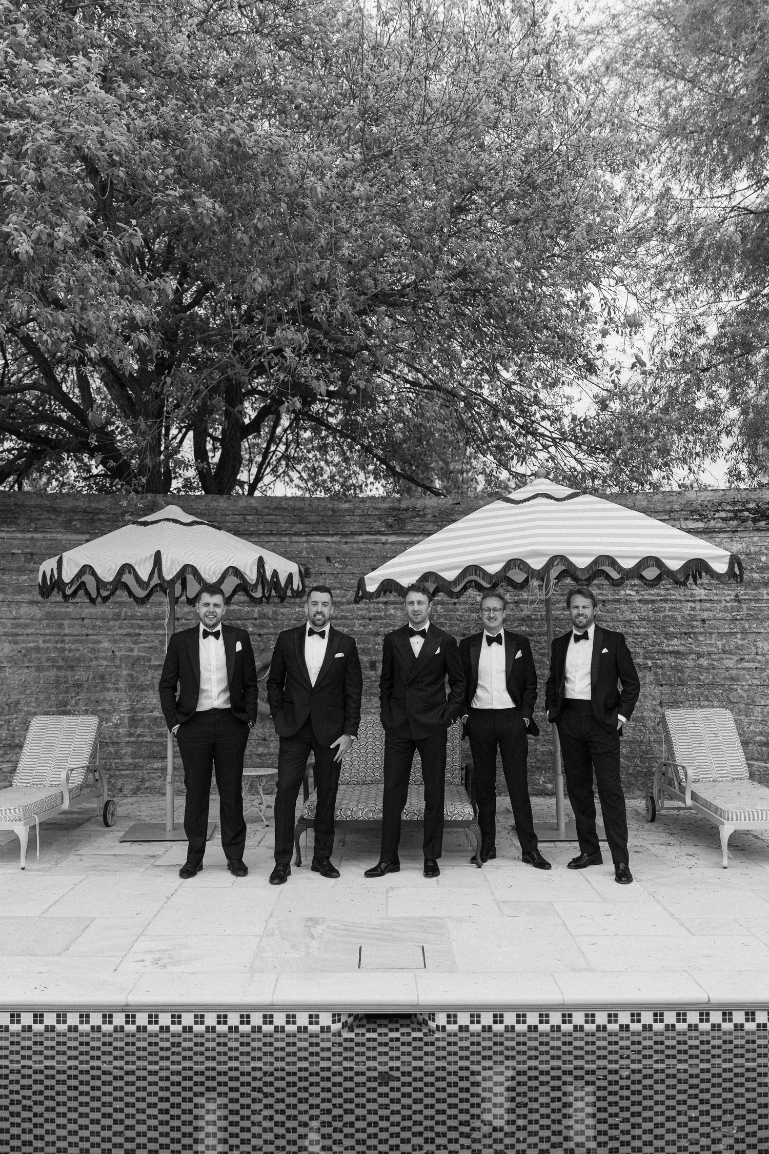 Five groomsmen in matching black tuxedos standing confidently by a poolside brick wall.