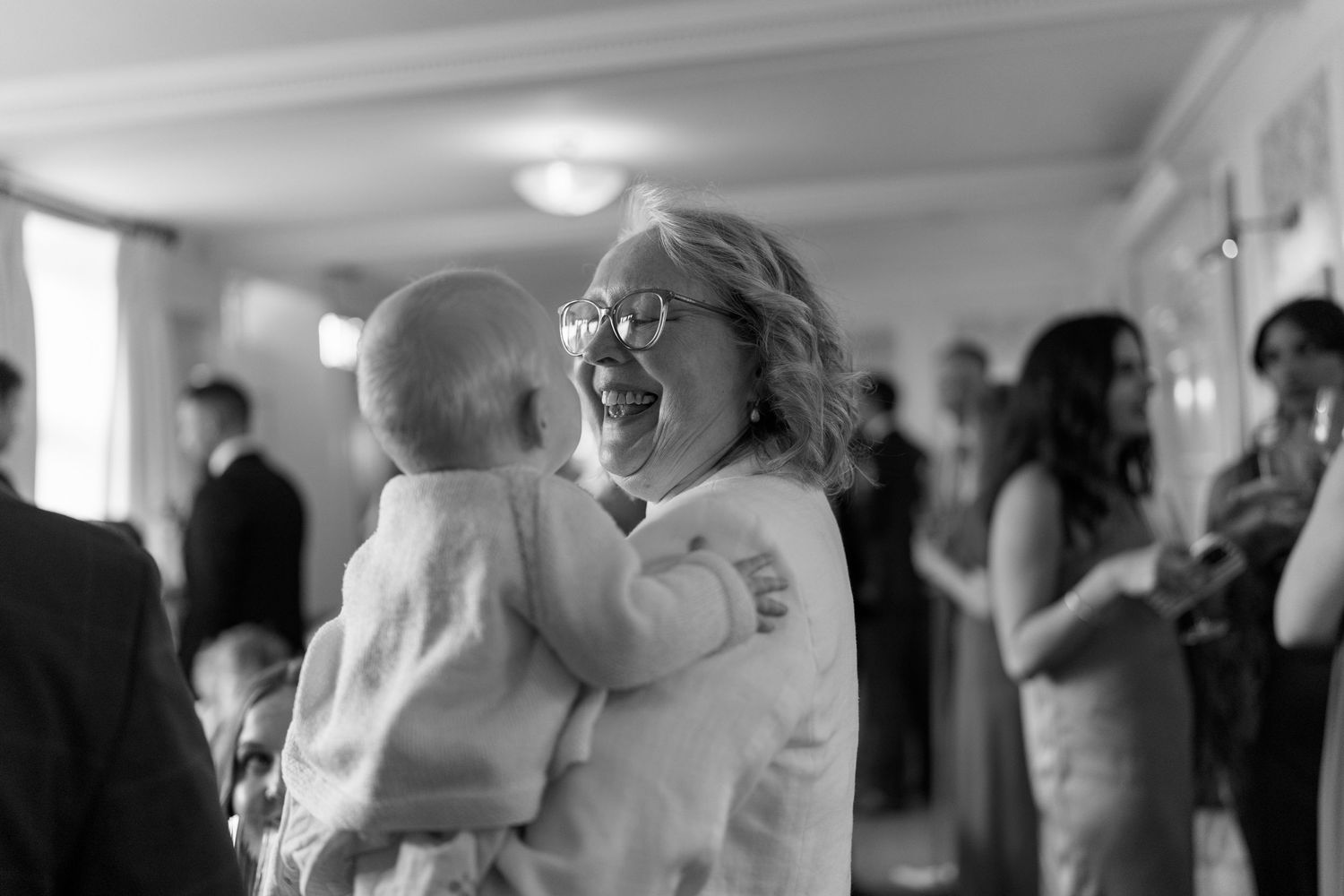 Joyful woman with glasses laughing while holding a baby at a wedding reception.
