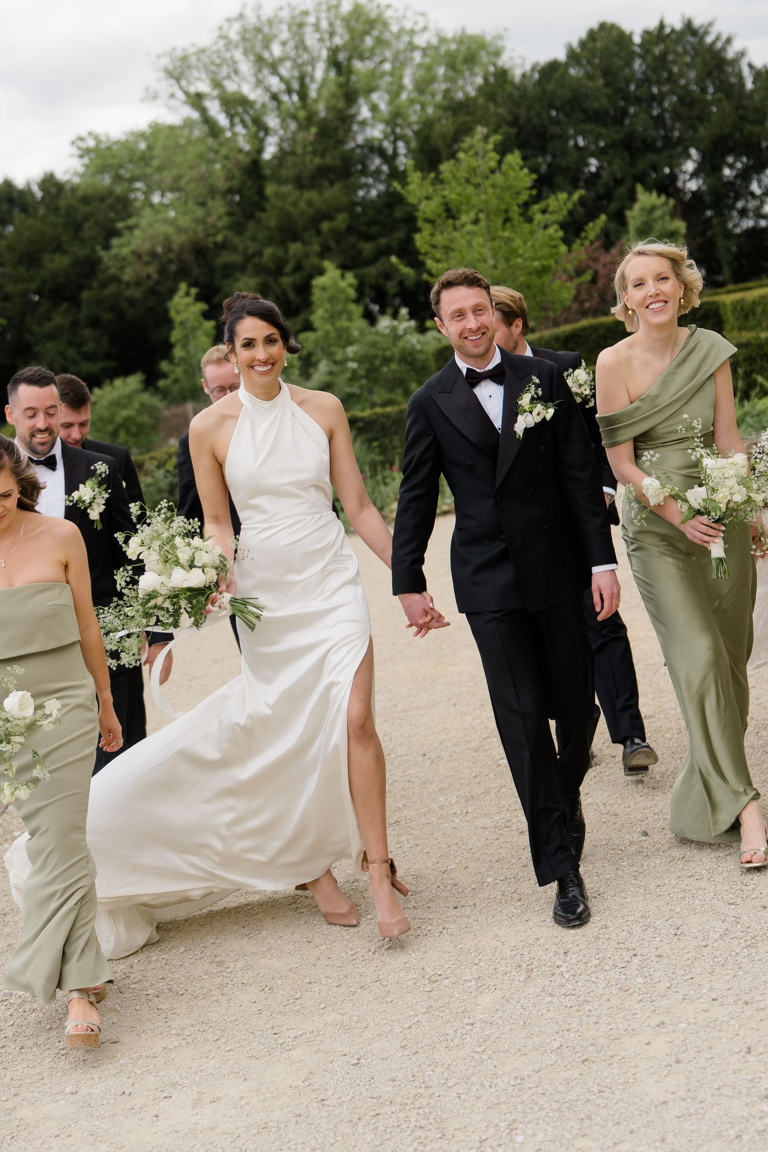 Bride and groom walk hand in hand with wedding party in sage green gowns along a gravel garden path.