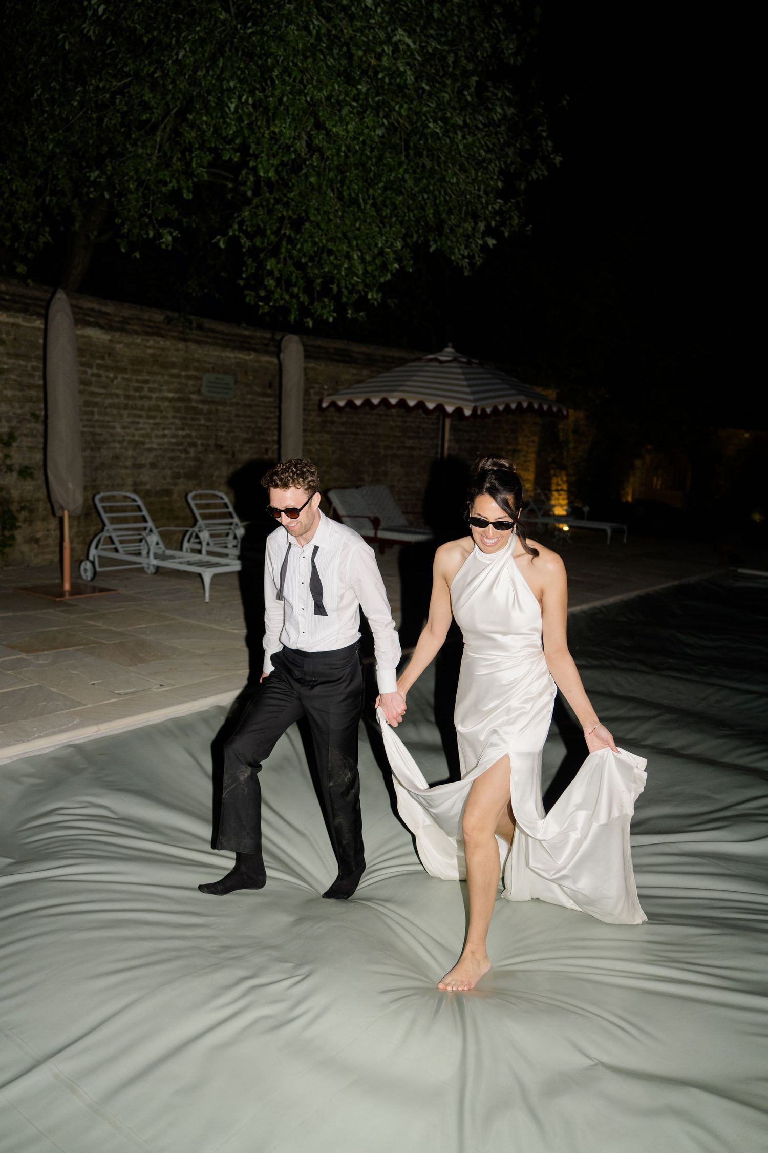 Elegant bride in white gown and groom in black suit stroll hand in hand by poolside at night.