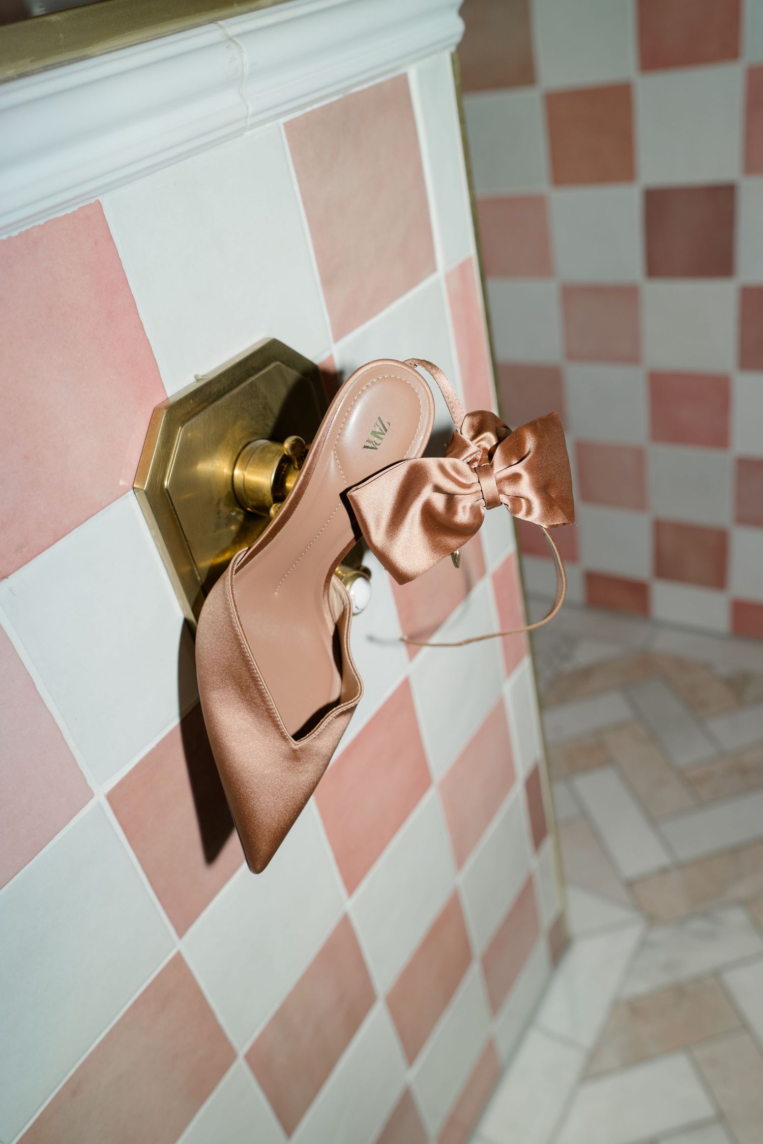 Blush satin bridal heels with bow detail hanging on a brass door knob against pink checkered tiles.