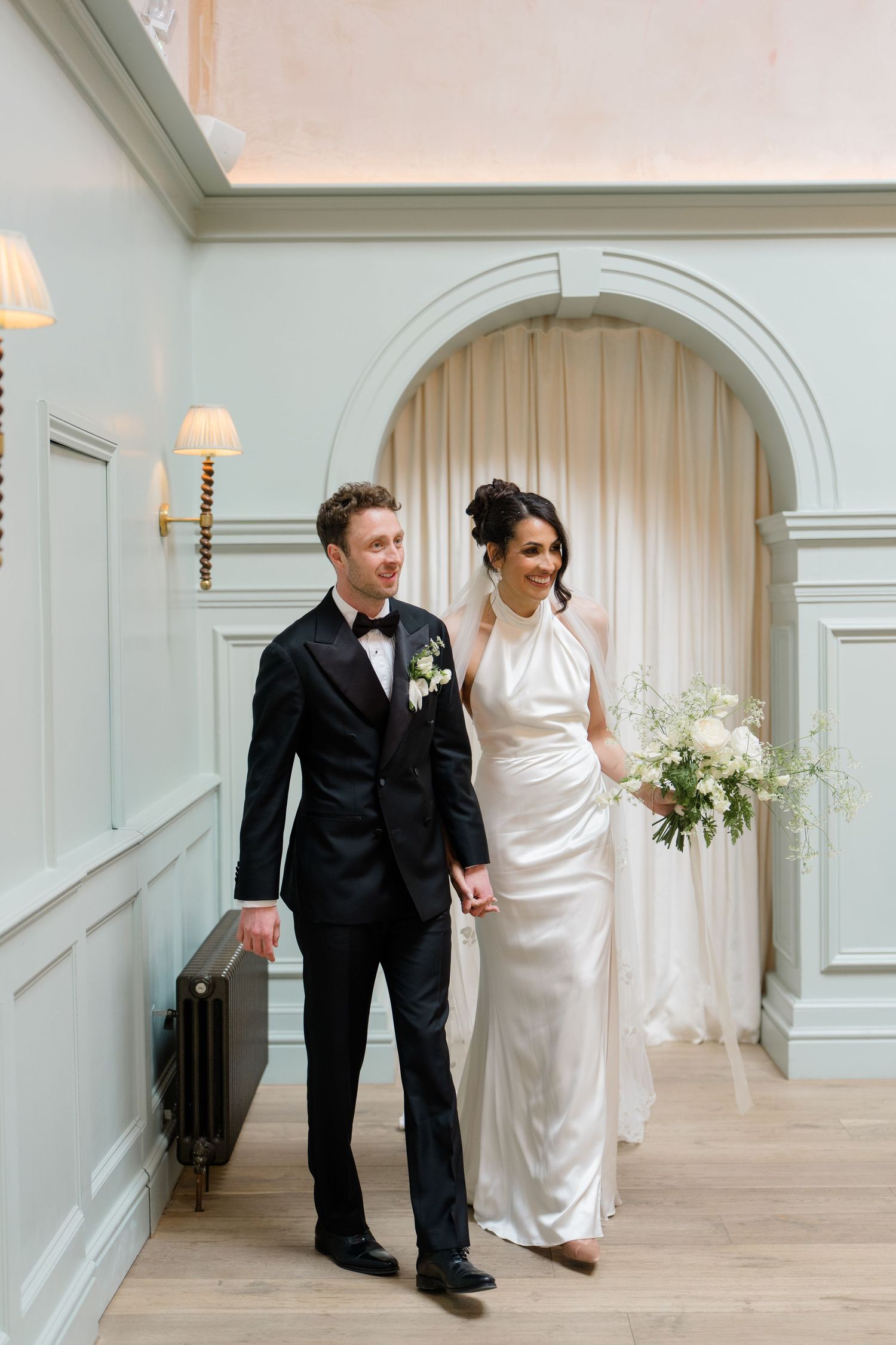 Elegant bride in halter neck satin gown and groom in black tuxedo walk hand in hand through a beautifully decorated wedding venue.