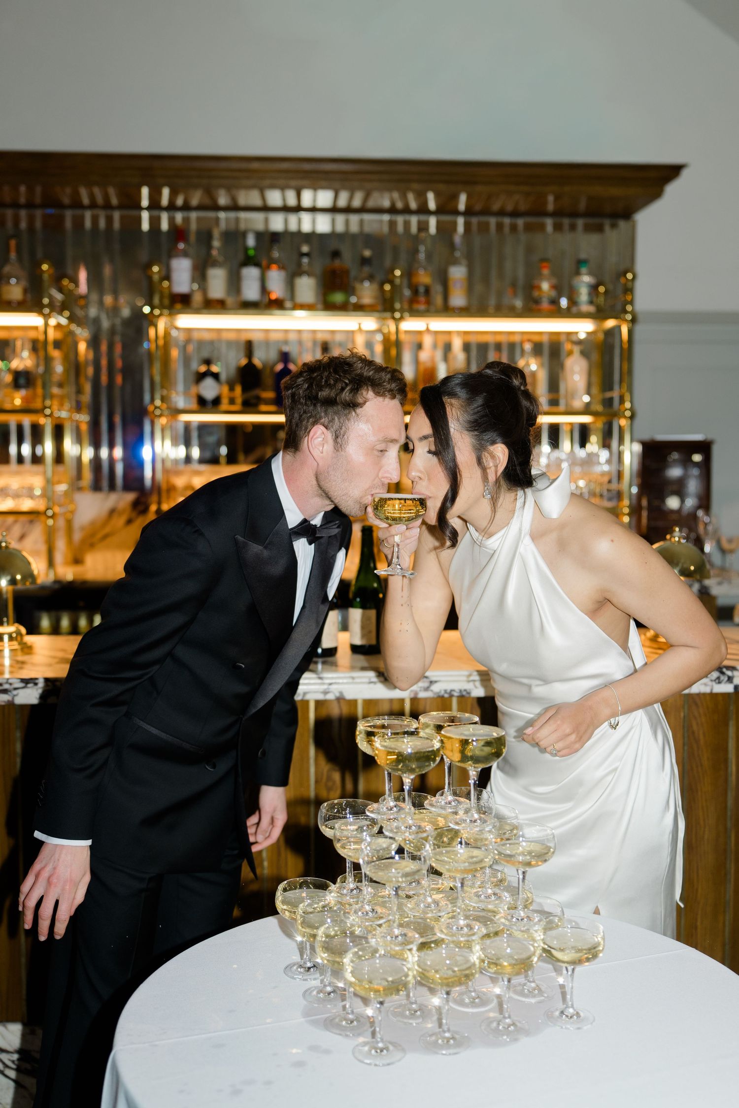 Bride and groom share champagne tower kiss at elegant wedding reception in front of gold bar.