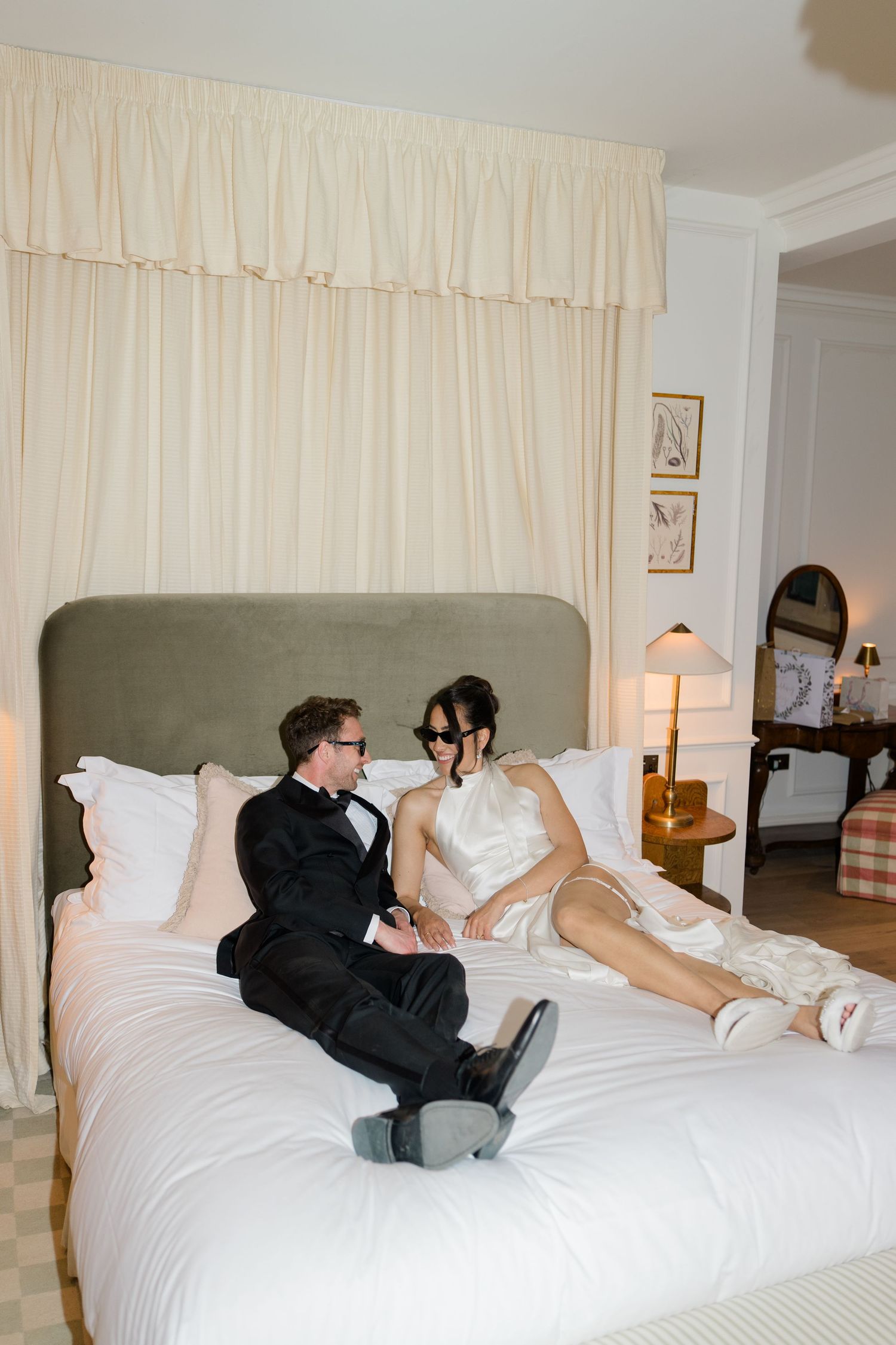 Bride and groom in wedding attire relaxing together on a luxurious white hotel bed.