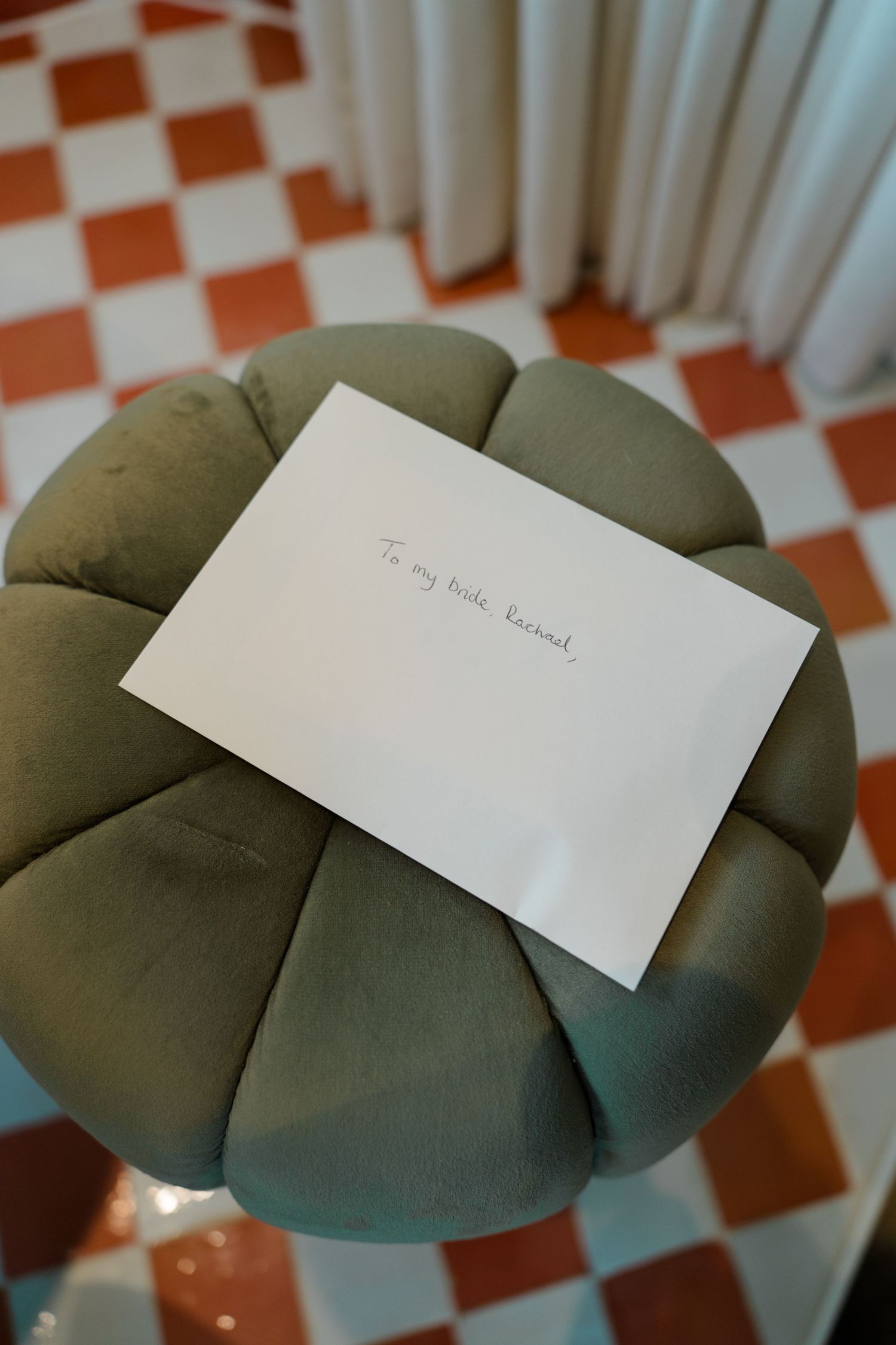 Handwritten wedding letter addressed to the bride resting on a green tufted ottoman with checkered floor.