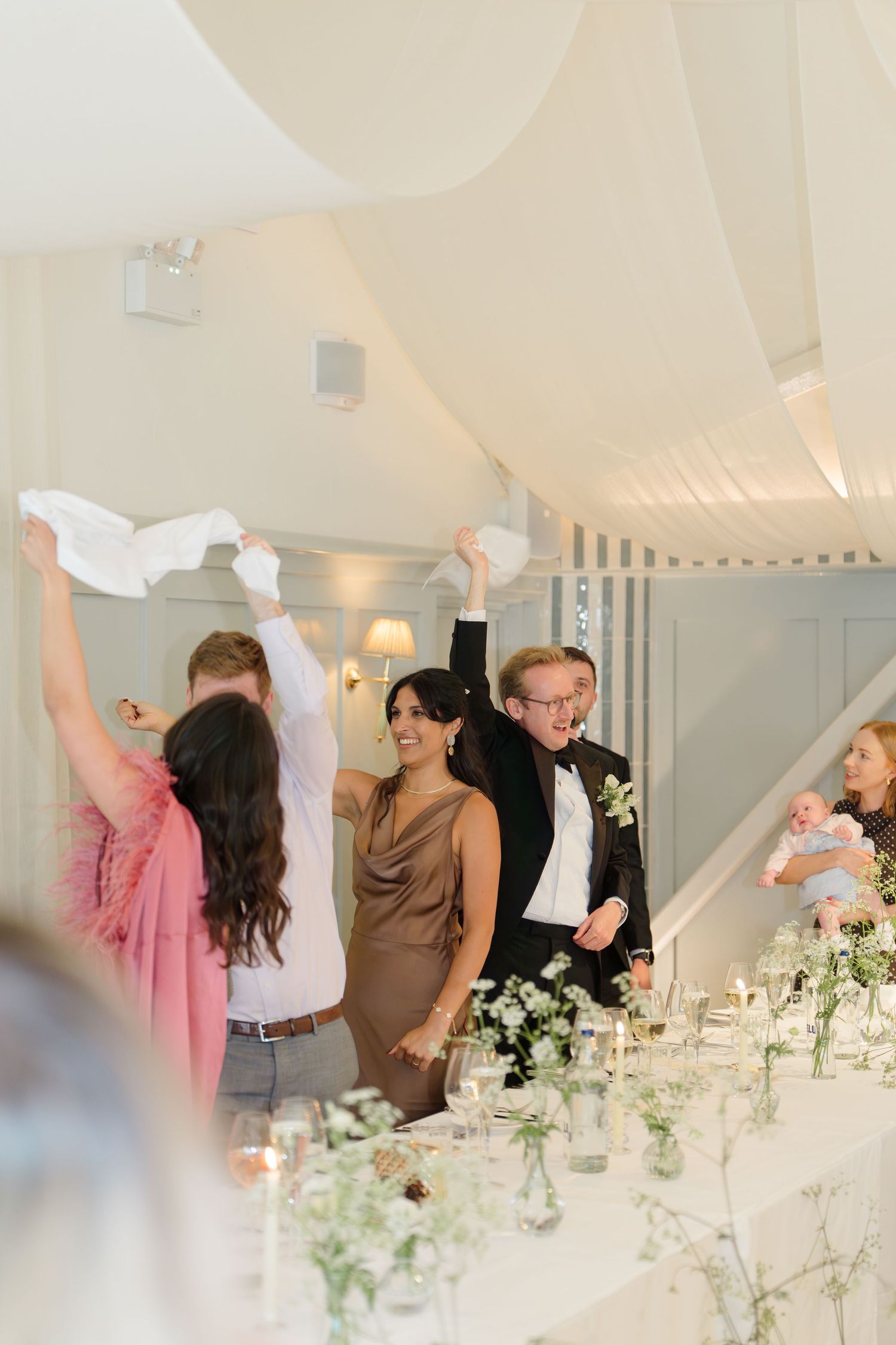 Joyful wedding guests waving white napkins in celebration at elegant white draped reception venue with floral table decor.