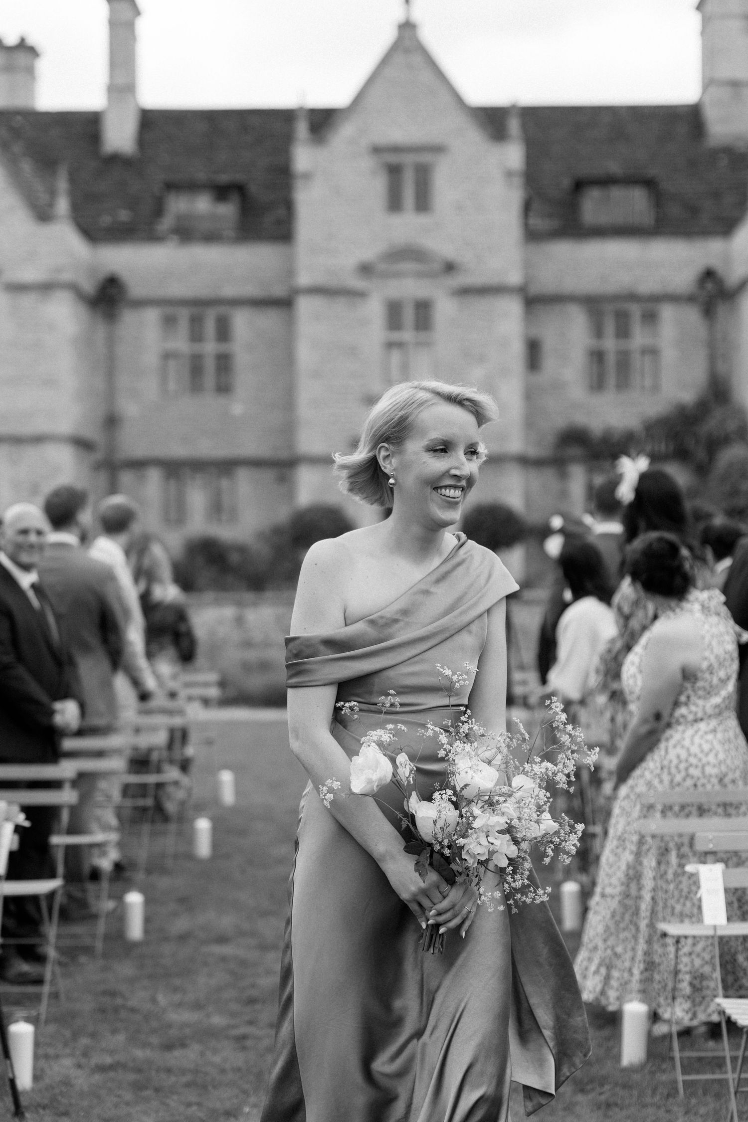 Smiling bridesmaid in gold gown carries bouquet outside historic manor wedding venue.