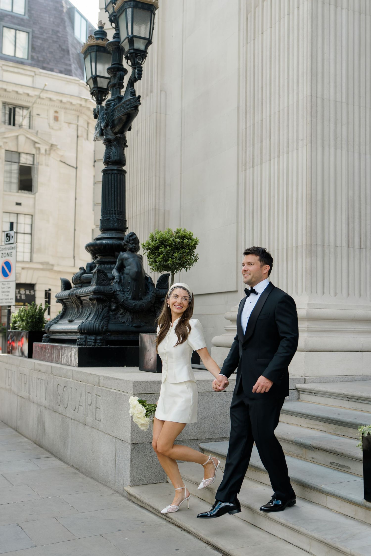 Stylish bride in white mini dress and groom in tuxedo walking hand in hand past ornate city lamppost.