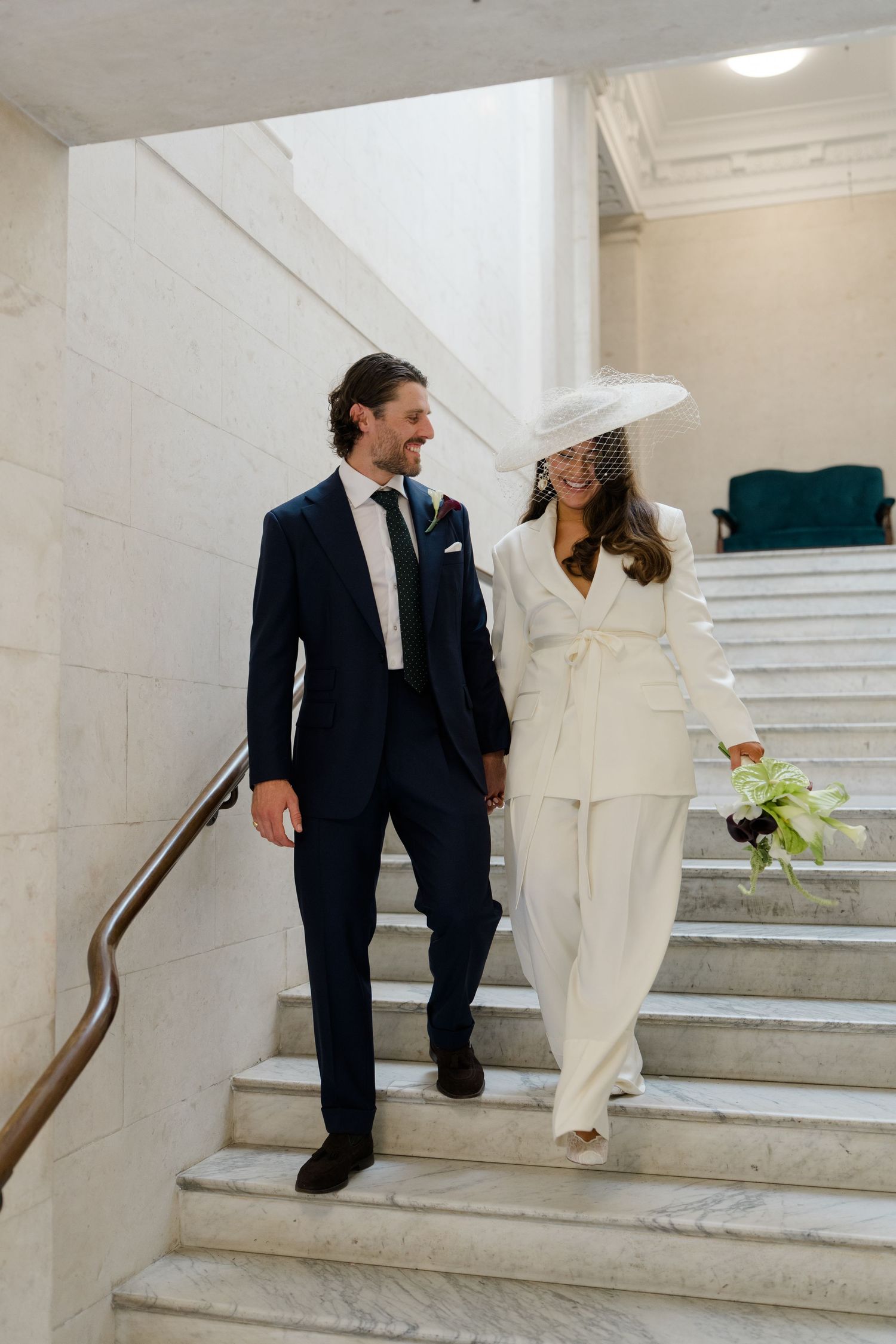 Couple in elegant wedding attire descending marble stairs, groom in navy suit, bride in white pantsuit and fascinator hat.