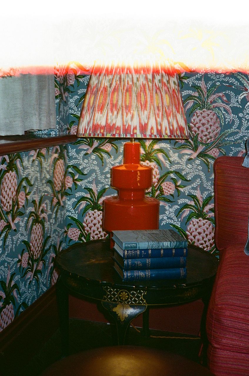 Orange ceramic lamp with ikat shade on dark side table with blue books against pineapple print wallpaper.