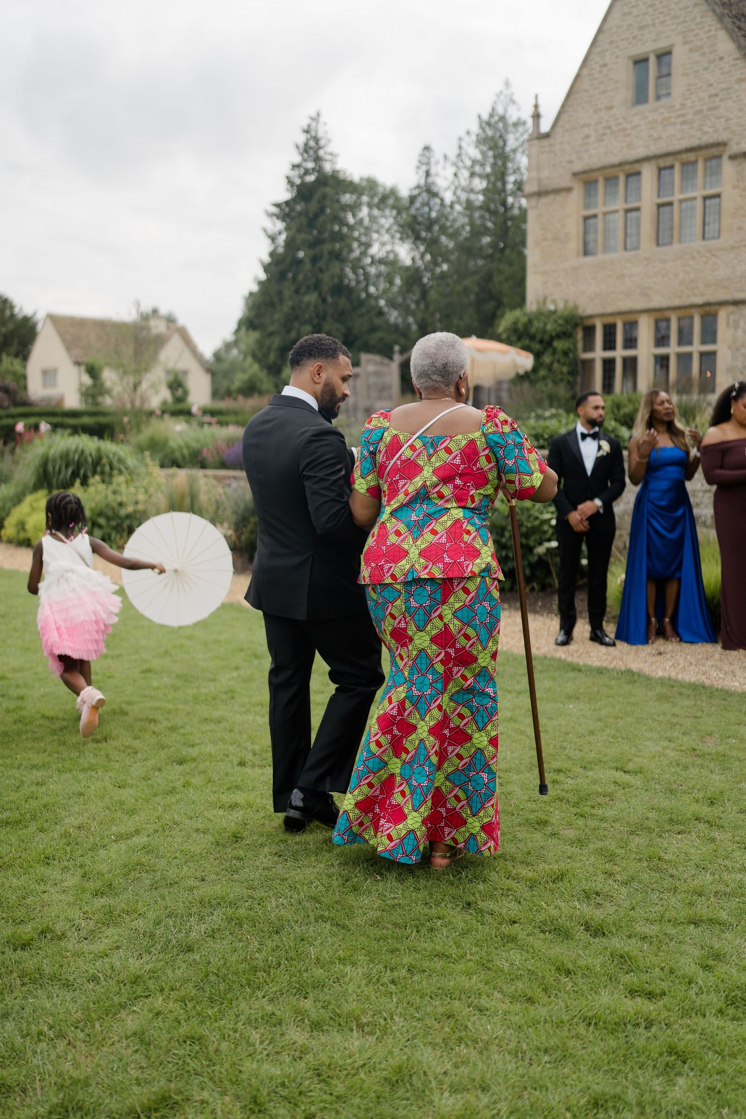 Groom escorts elderly woman in colorful African print dress across lawn at outdoor wedding reception.