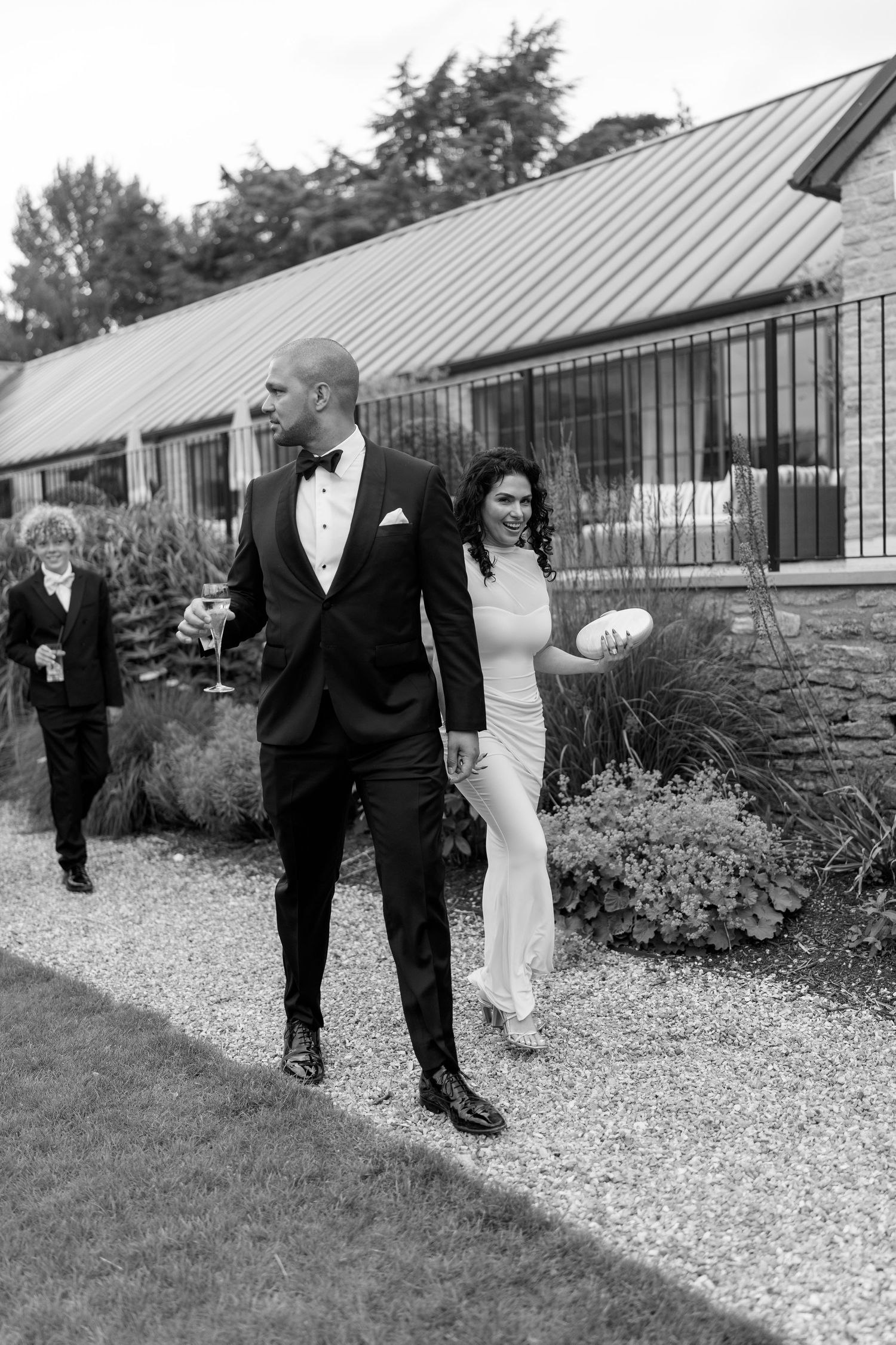 Groom in tuxedo walks with bride holding rugby ball on garden path.