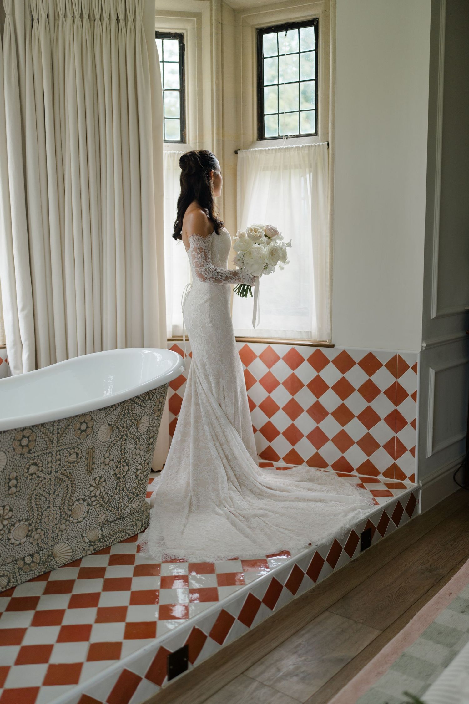 Bride in lace wedding gown with train holding white bouquet standing by window near clawfoot tub.
