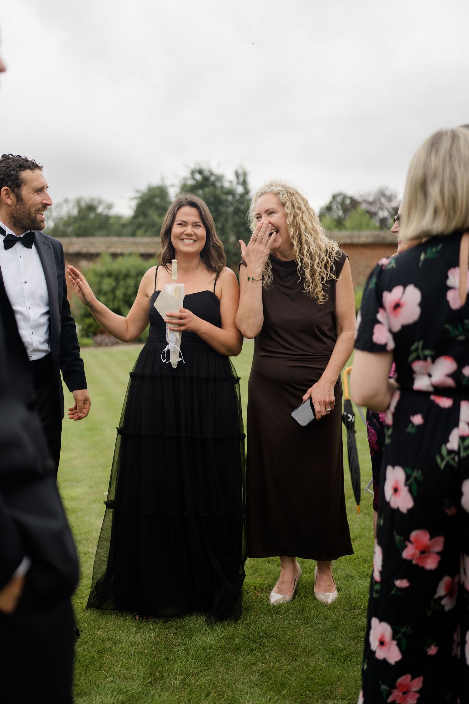 Wedding guests in black attire laughing and socializing on a green lawn at an outdoor reception.