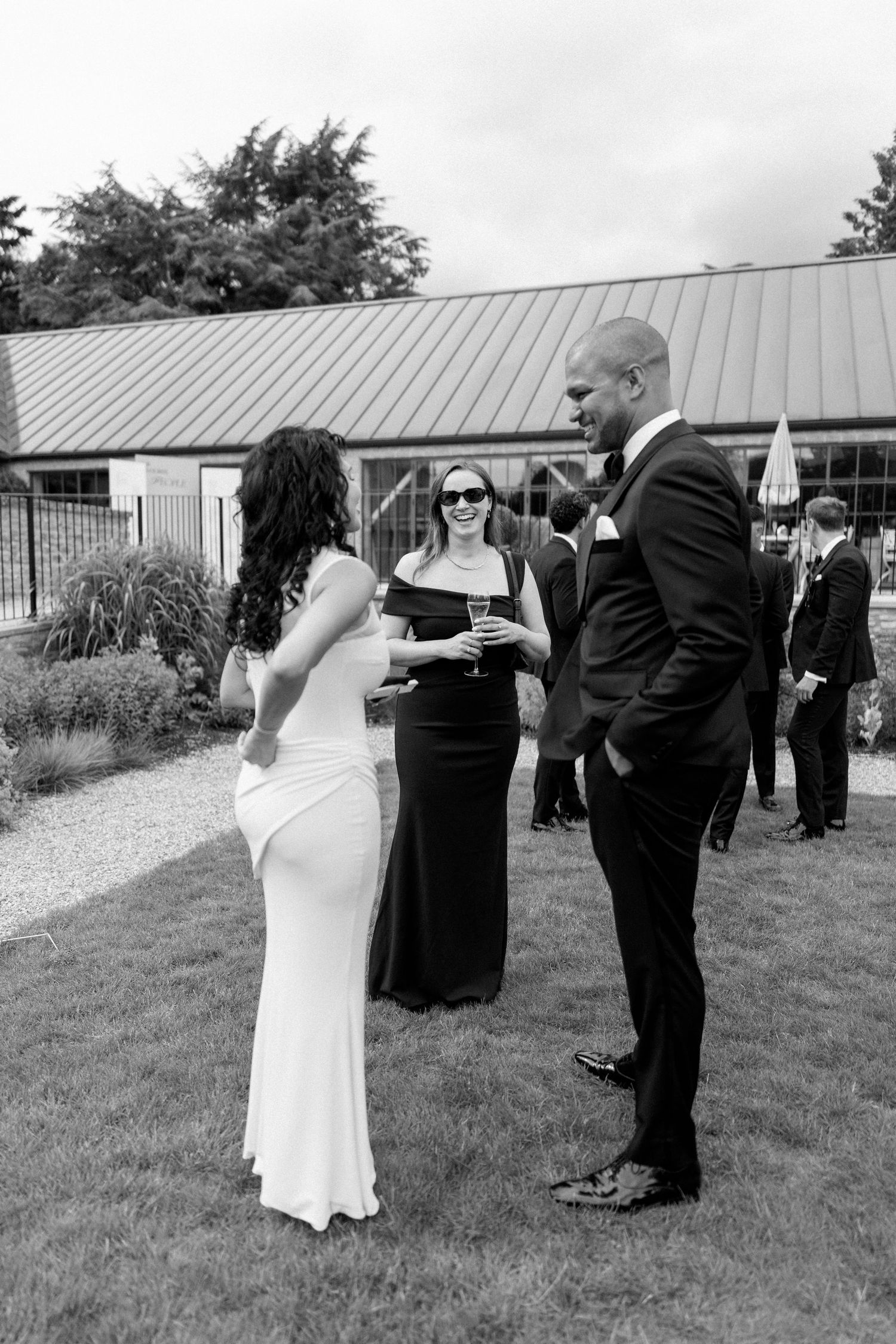 A black and white photo of a bride and guests conversing outdoors at a wedding reception venue.