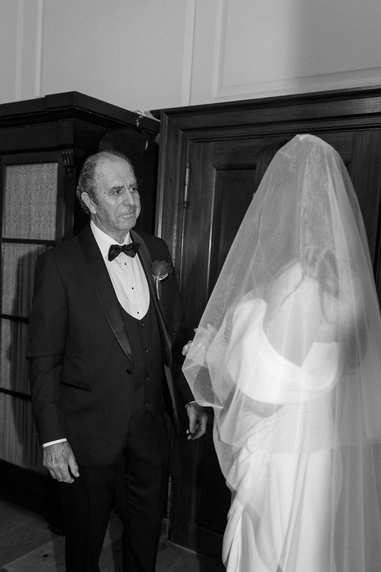 Black and white photo of a father in a tuxedo and bow tie watching a bride in a long sheer veil and gown before her wedding ceremony.