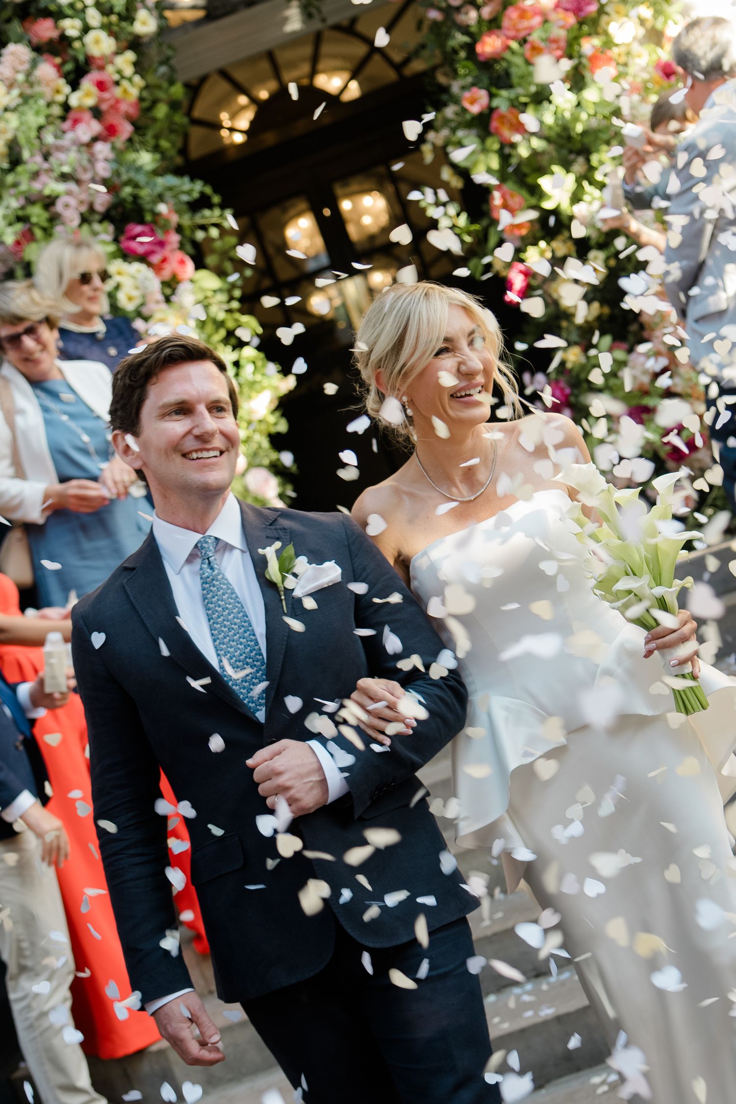 Joyful newlyweds walk through confetti shower, bride holding white bouquet, groom in navy suit, floral arch backdrop.