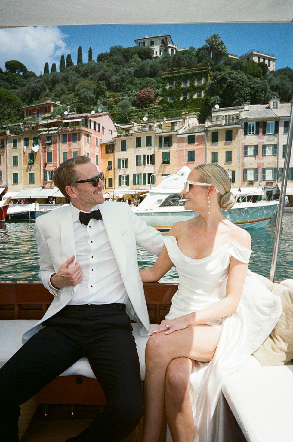 Elegant couple in wedding attire sitting on a boat in Portofino, Italy, with colorful harbor buildings in the background.