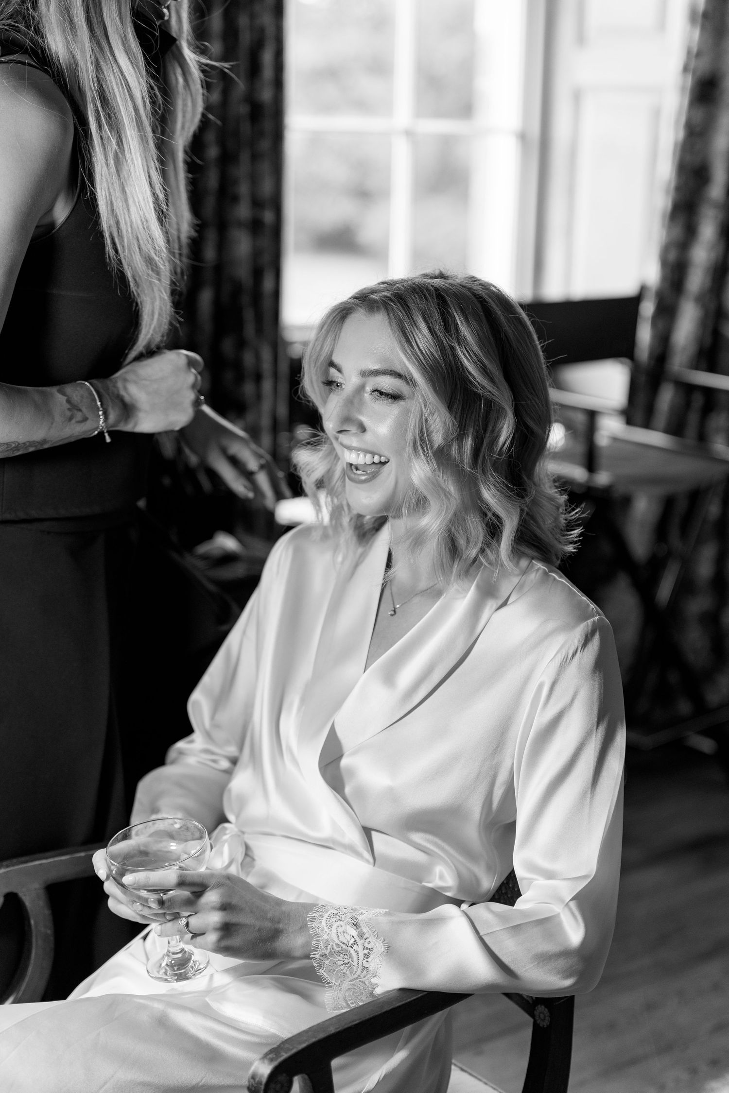 Bride laughing in white satin robe holding champagne glass during wedding preparations.