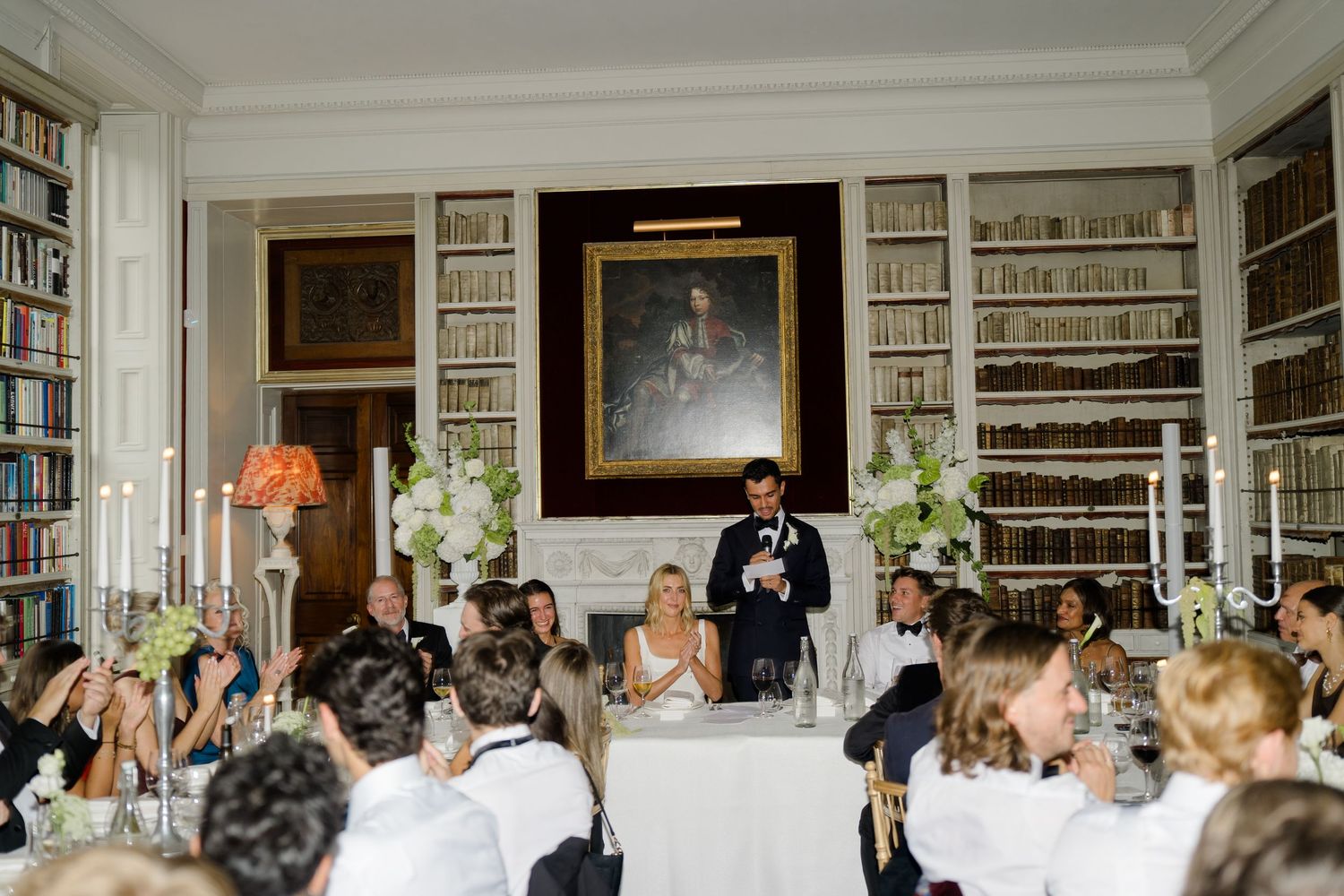 A groom delivers a wedding speech in a grand library with guests raising glasses.