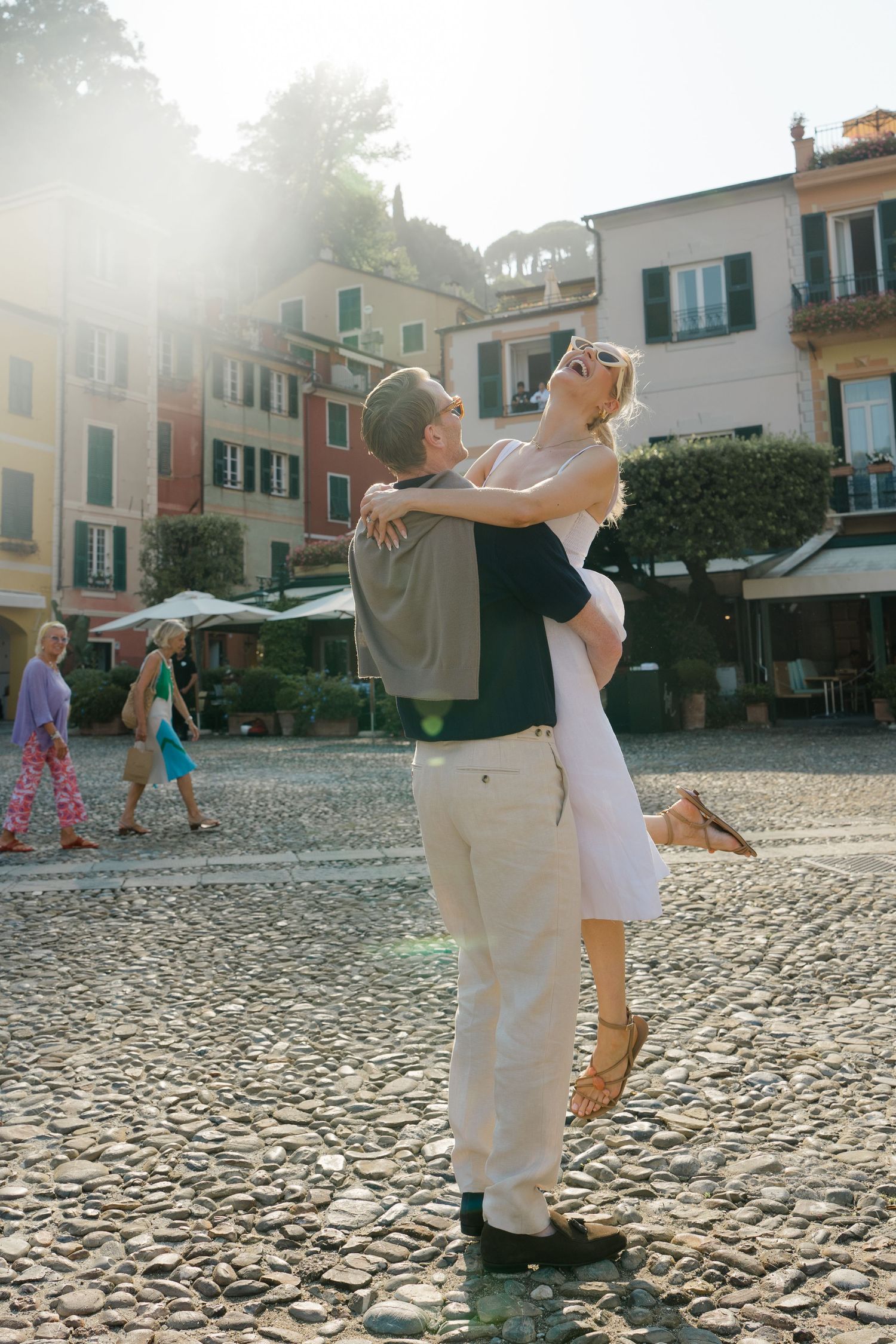 Couple embracing and dancing in a sunlit cobblestone square surrounded by colorful Italian buildings.