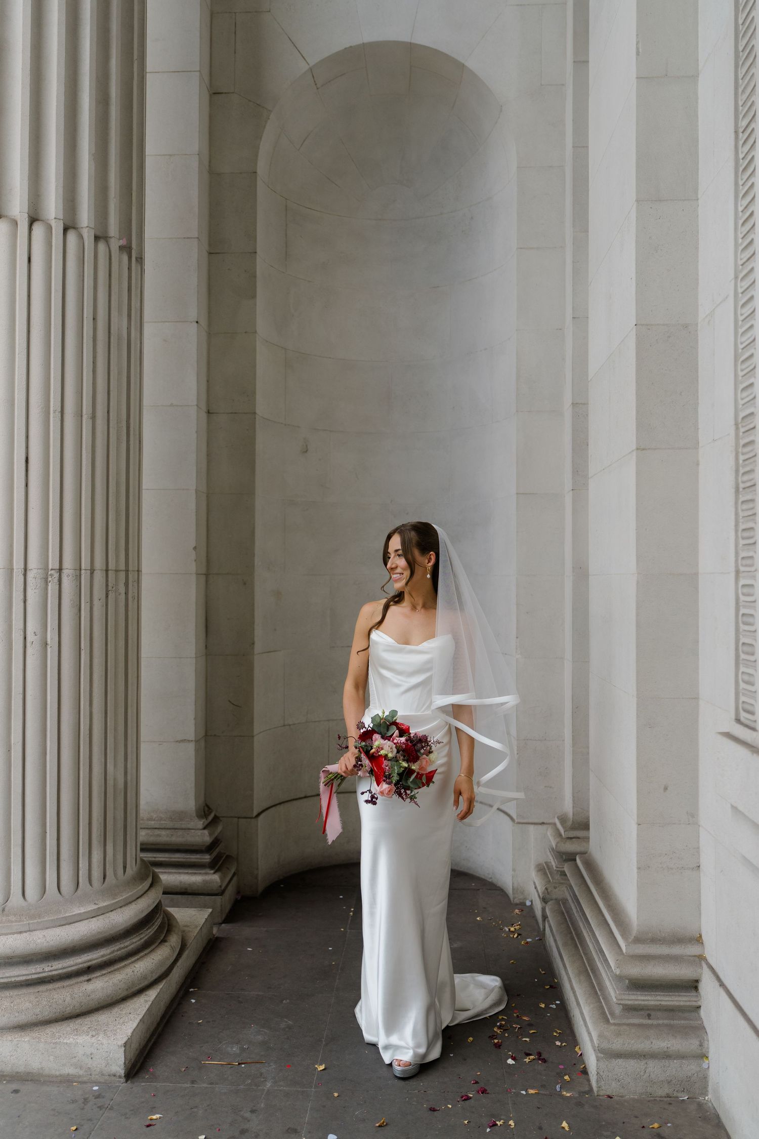 Bride in white satin gown and veil holding red bouquet between stone columns.