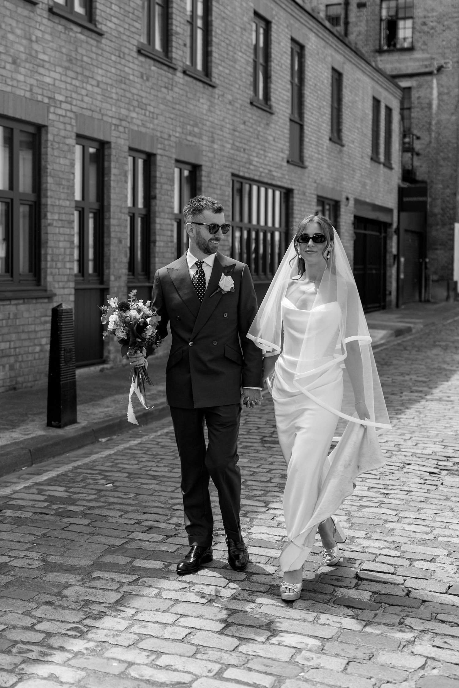 Bride and groom in sunglasses walk hand-in-hand on cobblestone street past brick buildings on their wedding day.