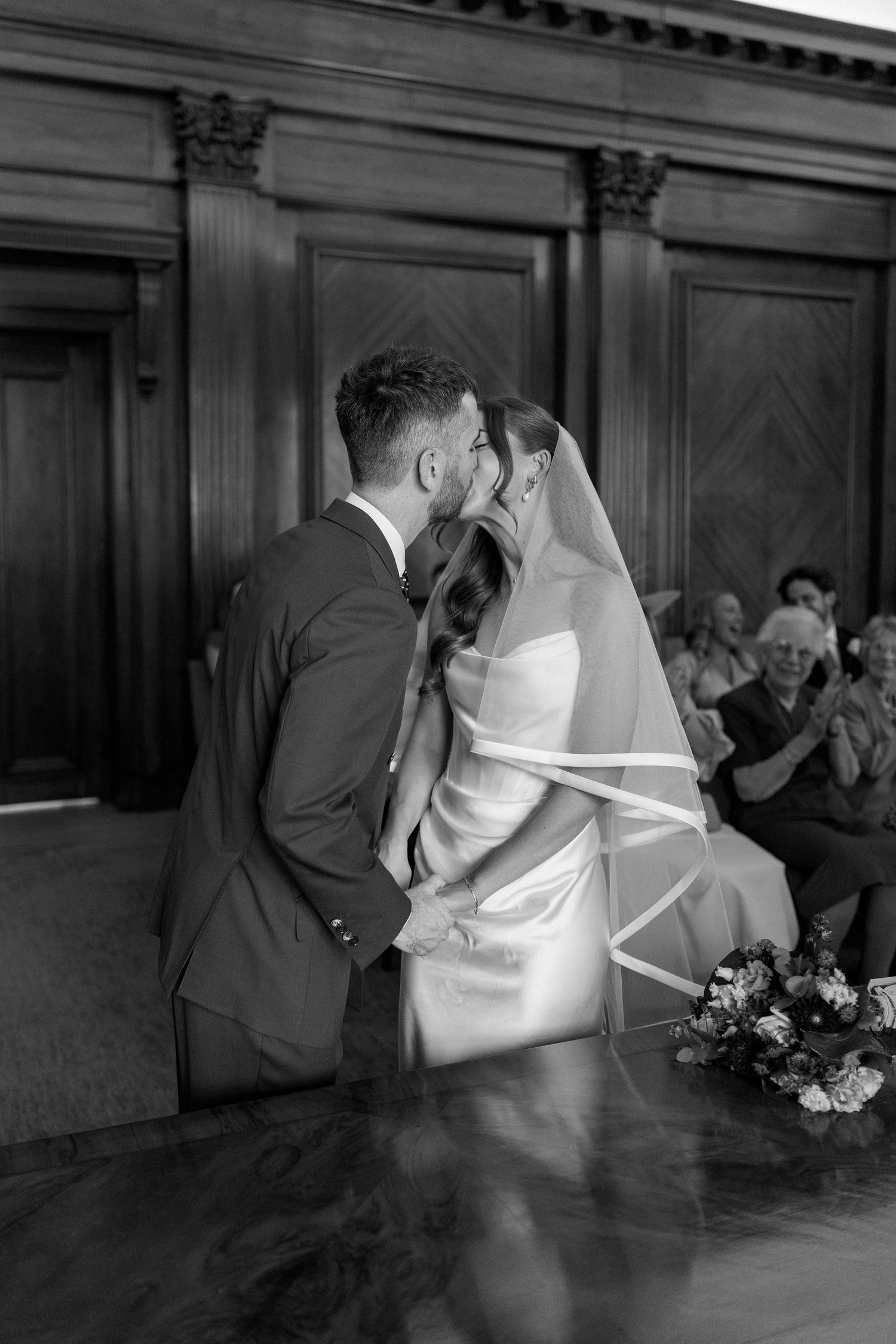 Bride and groom share first kiss at wedding ceremony in elegant wood-paneled venue, black and white.