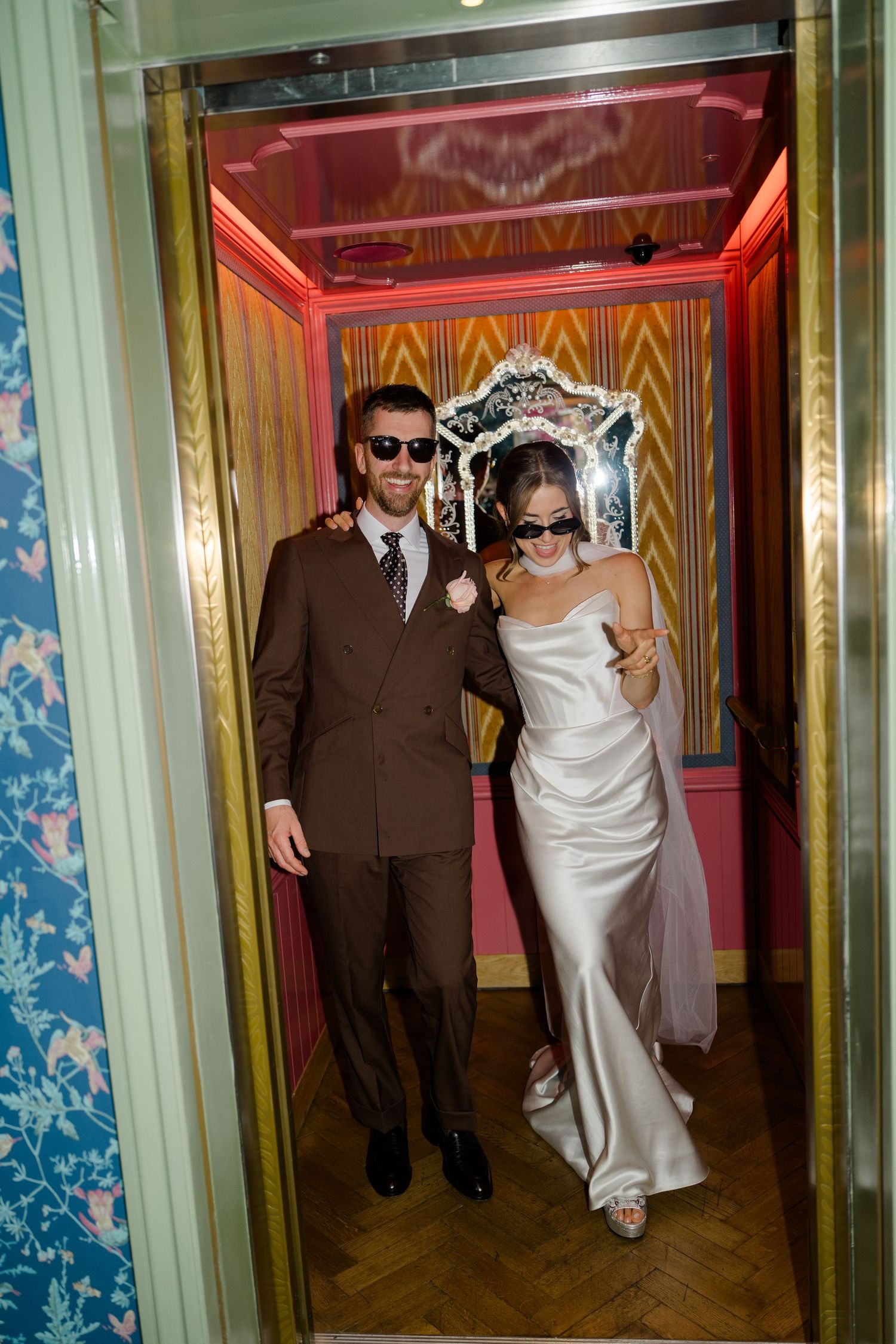 Bride and groom in sunglasses stepping out of ornate elevator in wedding attire.