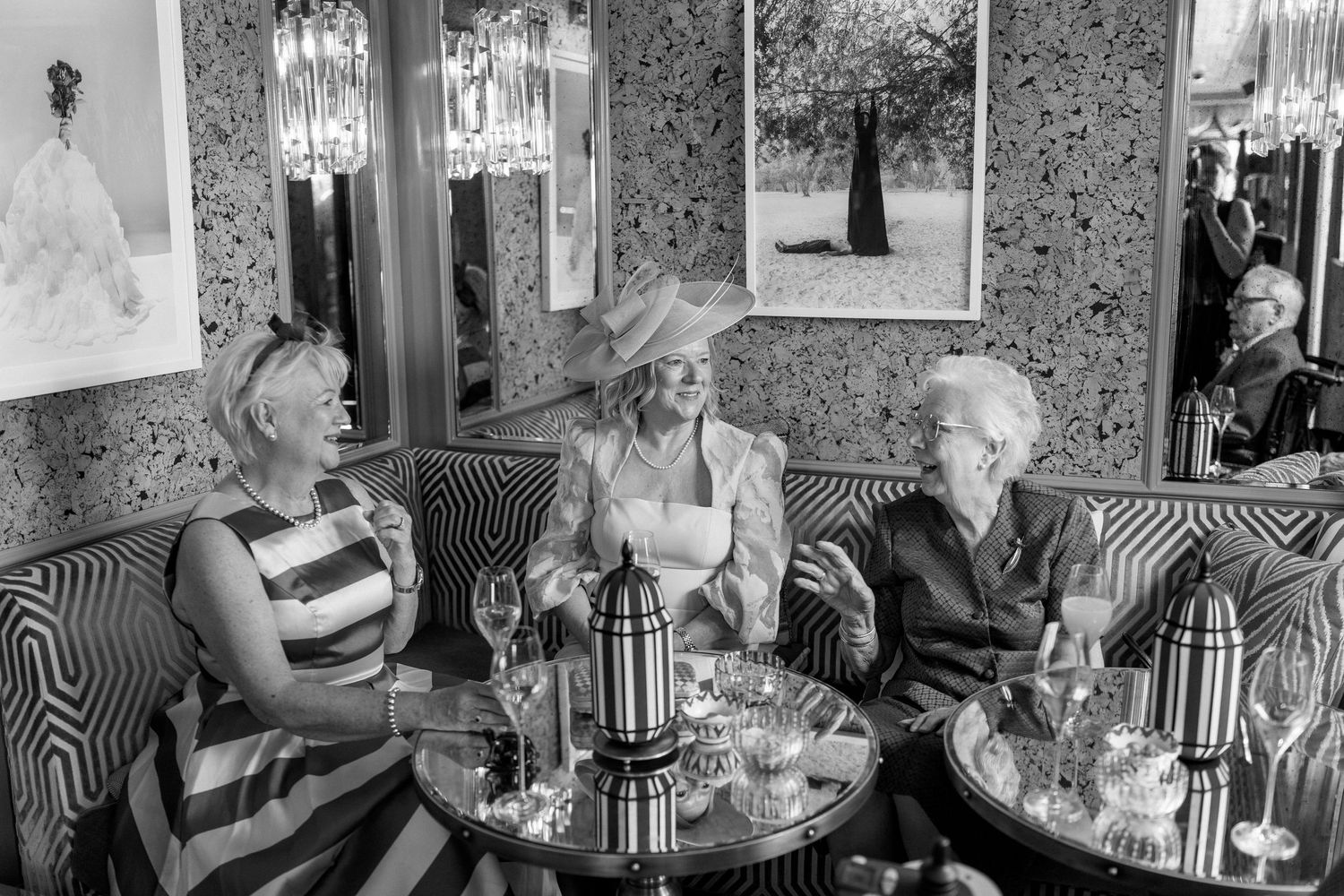 Three elegant women laugh together at a stylish restaurant booth during a wedding celebration in black and white.