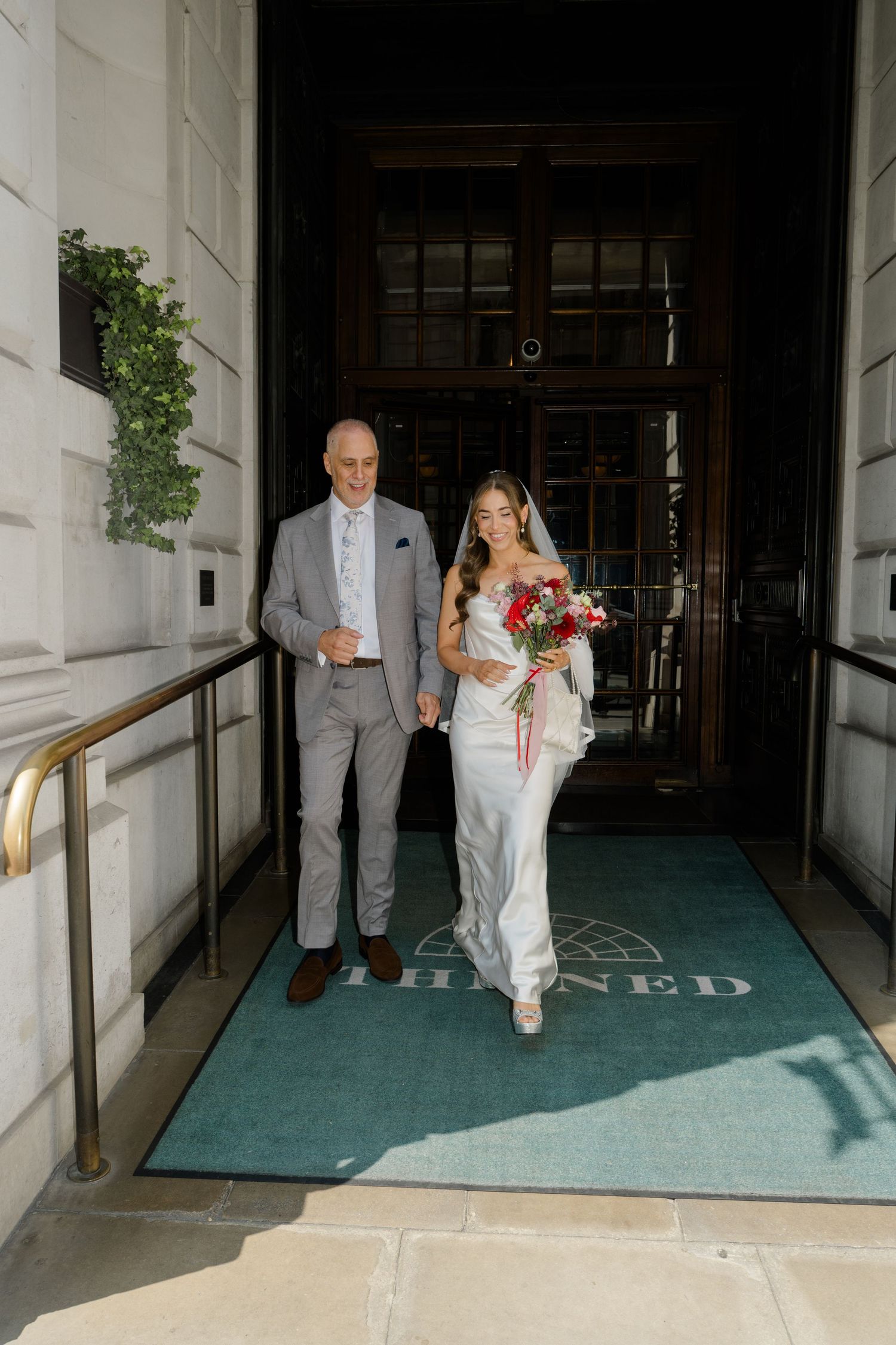 Bride in white gown carrying red bouquet walks with older man from The Ned hotel entrance.
