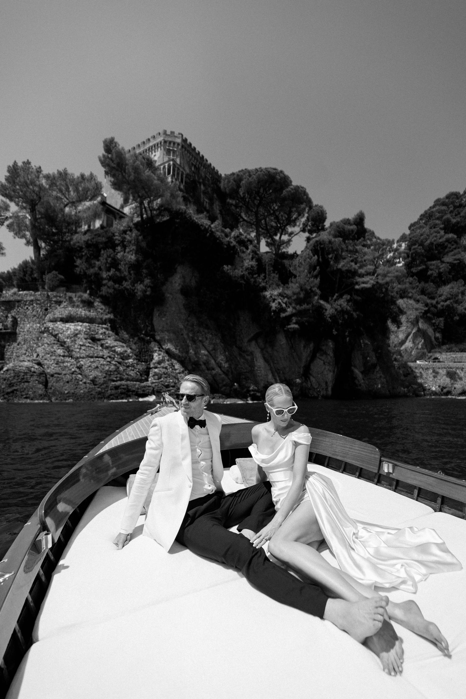 Bride and groom relaxing on luxury boat bow with dramatic rocky coastline behind them.