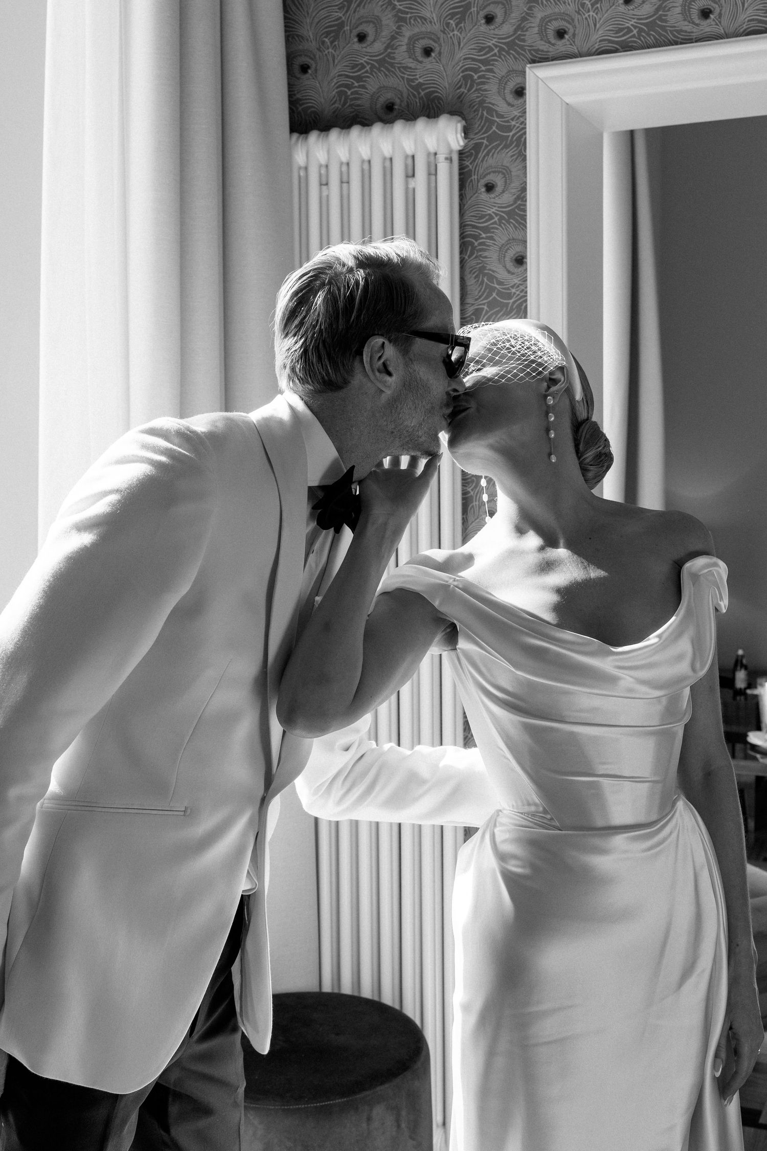 Black and white photo of bride and groom sharing a tender kiss by a sunlit window indoors.