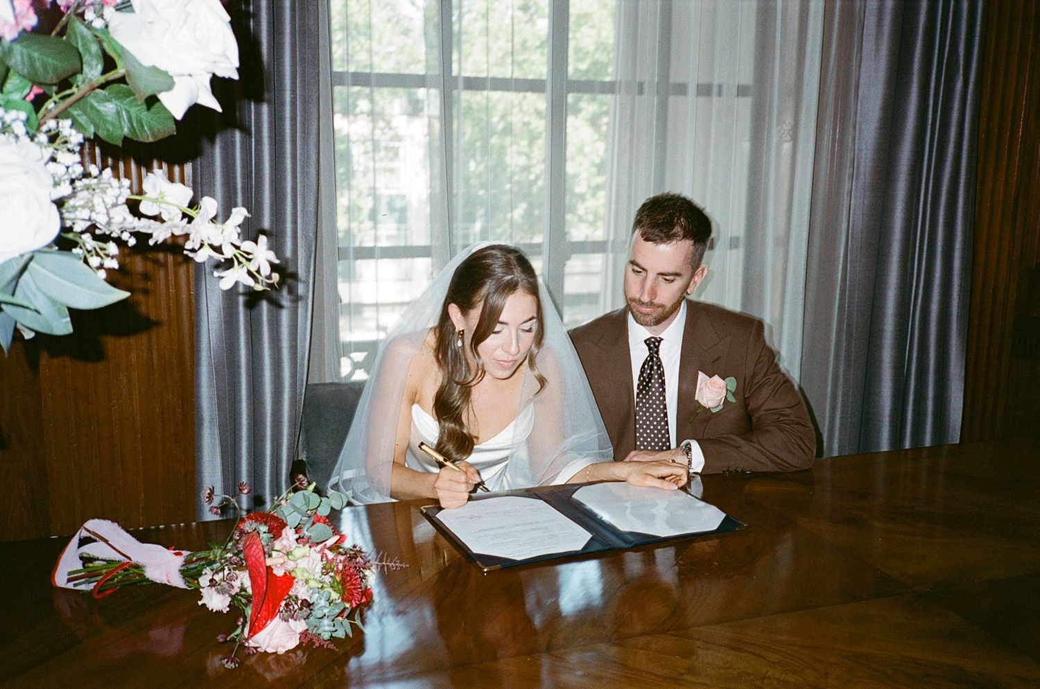 Bride and groom signing wedding documents at a wooden table with a colorful bouquet nearby.