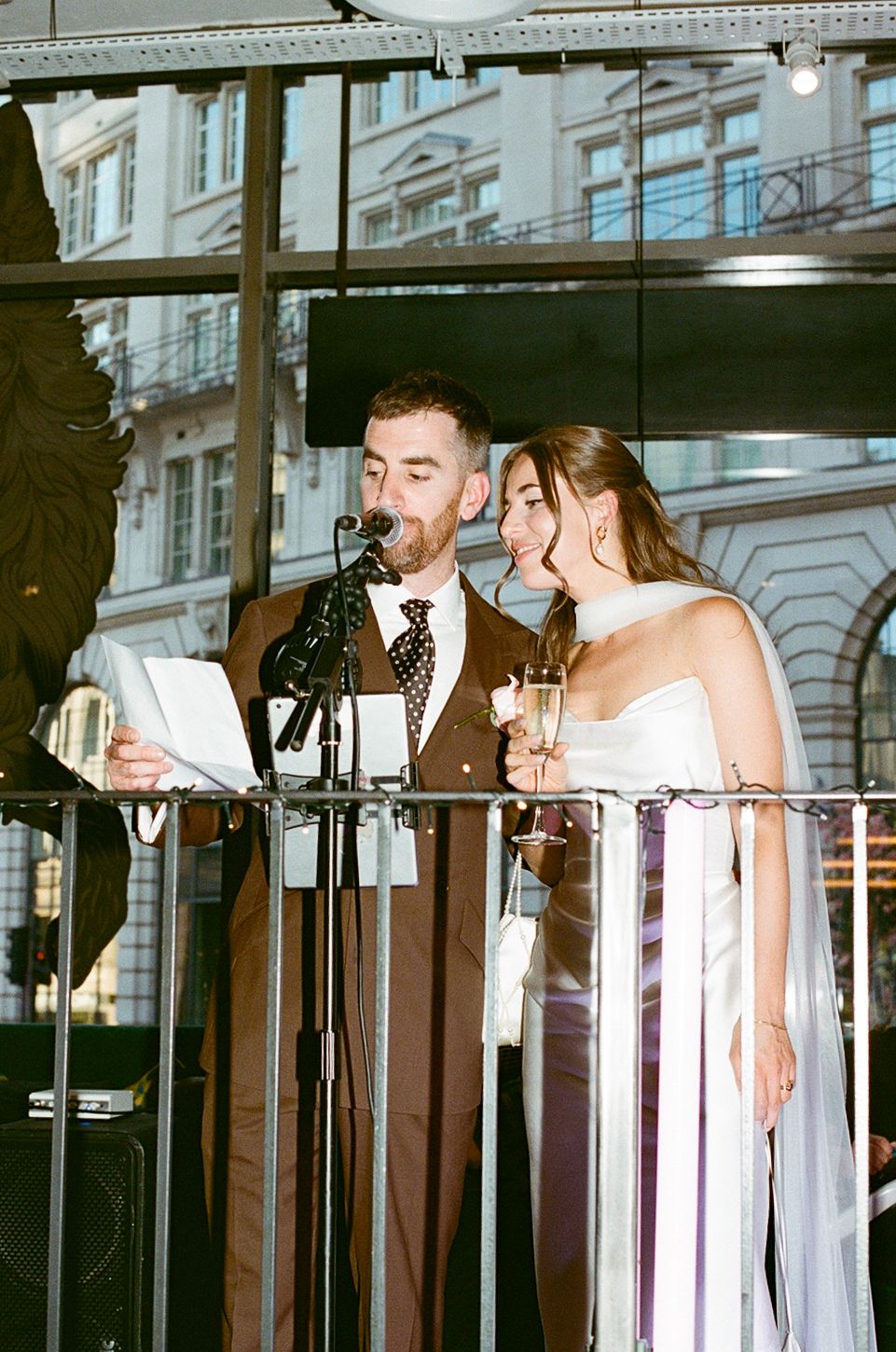 Bride and groom giving speech together at microphone on balcony at evening reception.