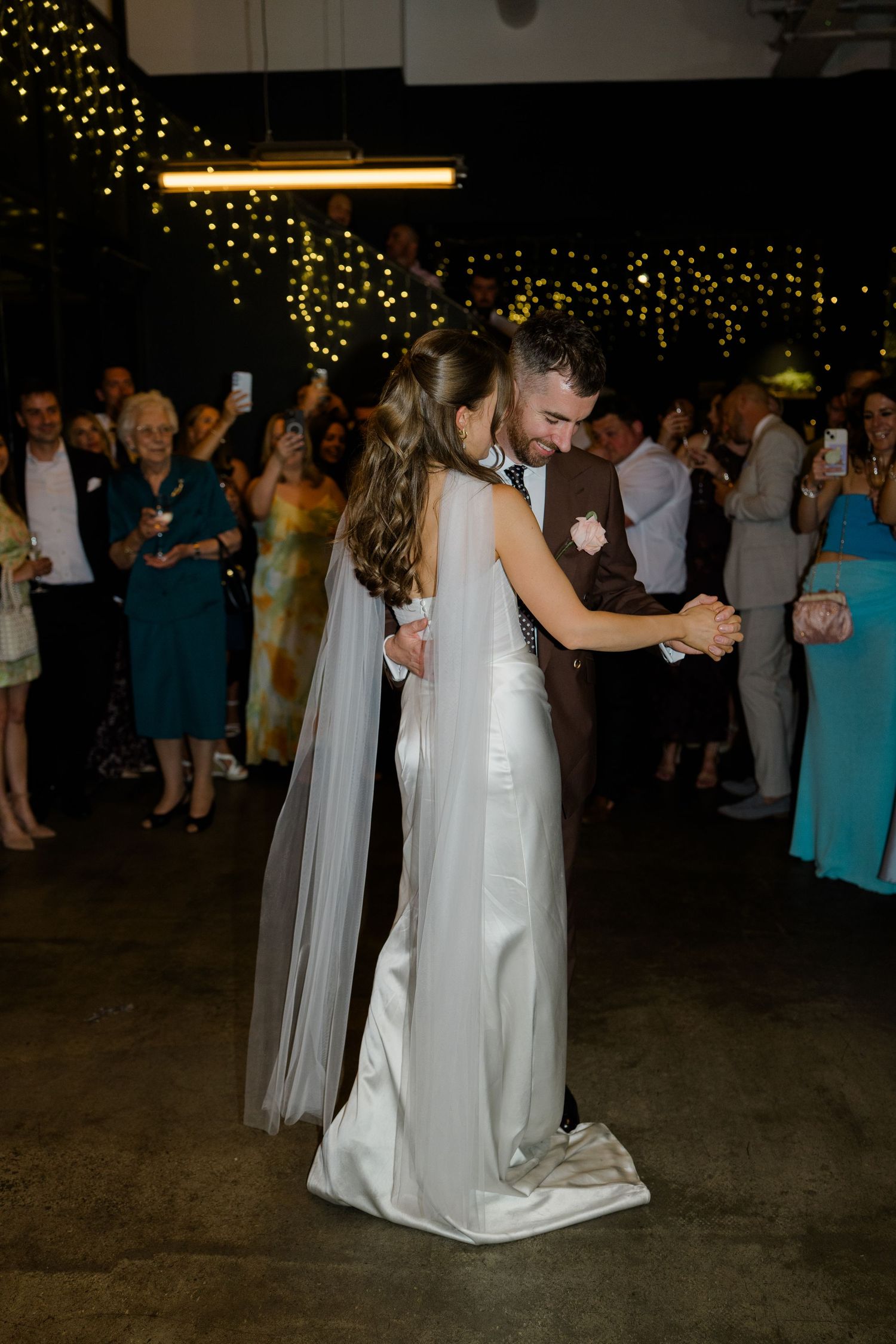 Bride and groom share first dance surrounded by guests with fairy lights backdrop.