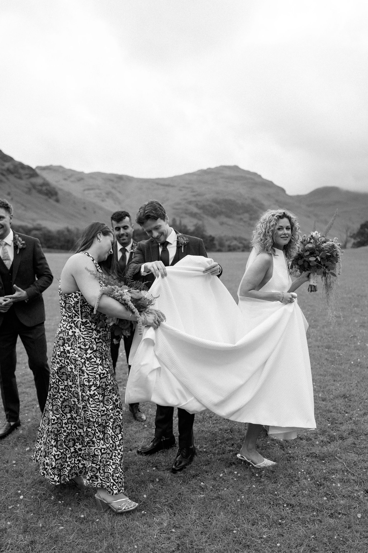 Wedding party helping with the bride's dress in a playful moment against mountain scenery.