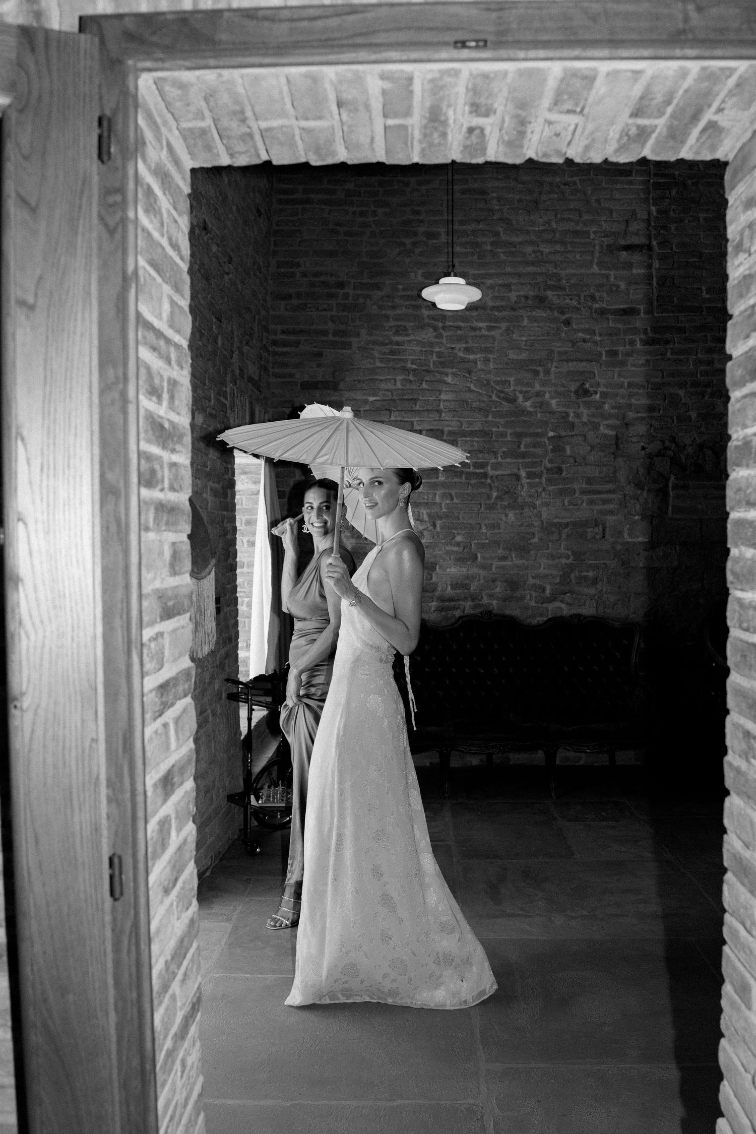 Figure in white dress holding parasol poses dramatically in dark brick interior space.