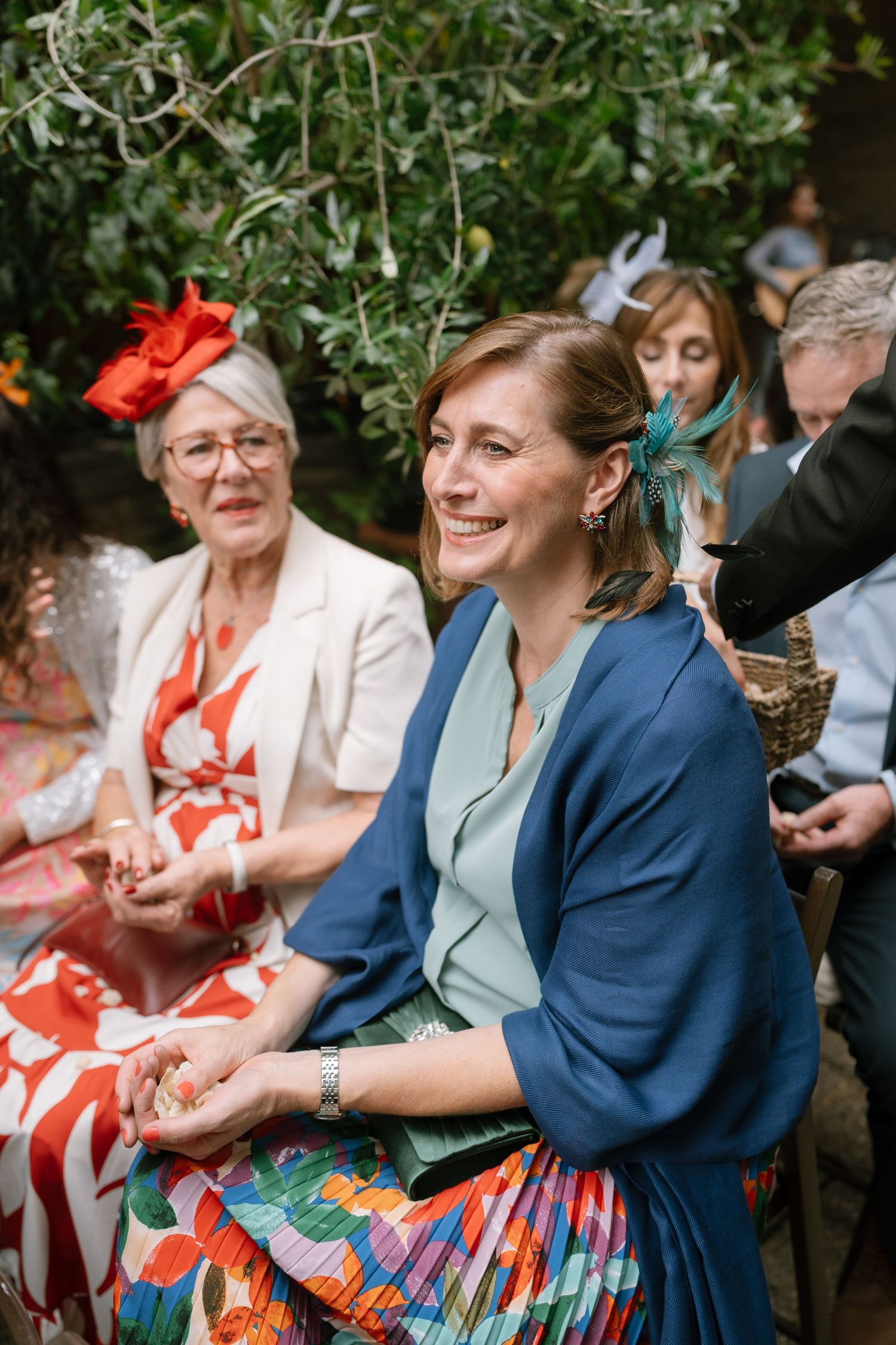 Wedding guests in colorful attire seated outdoors under trees during a celebration.