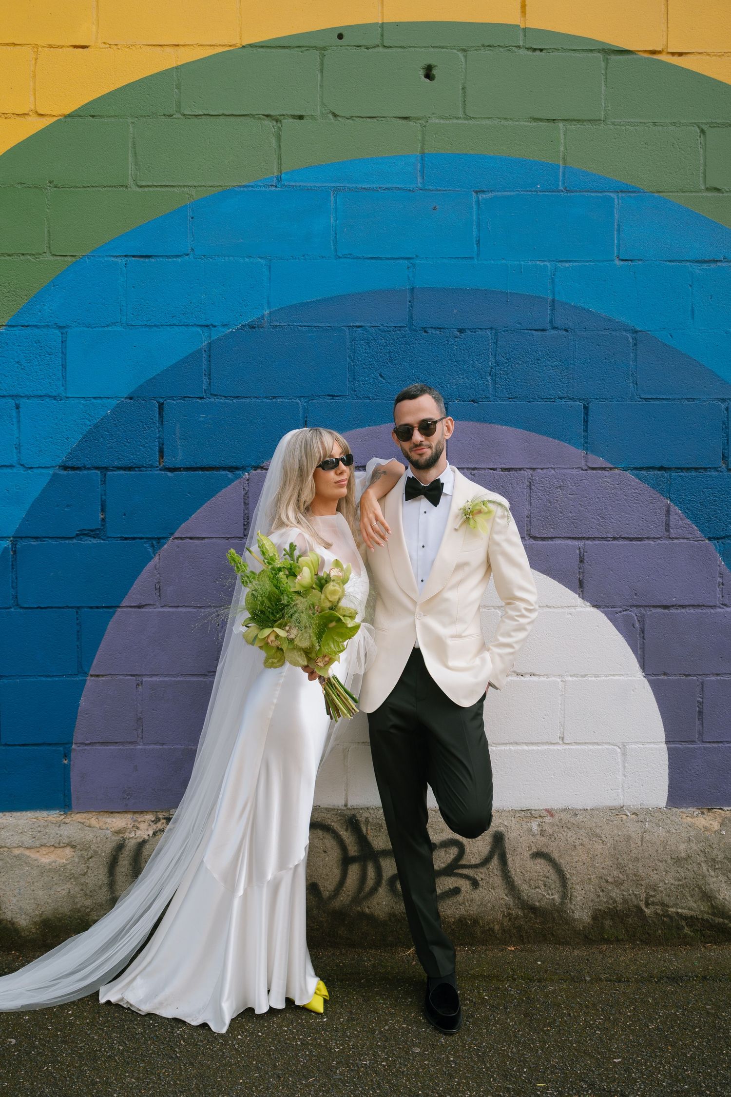 A wedding couple poses against a colorful rainbow painted brick wall backdrop.