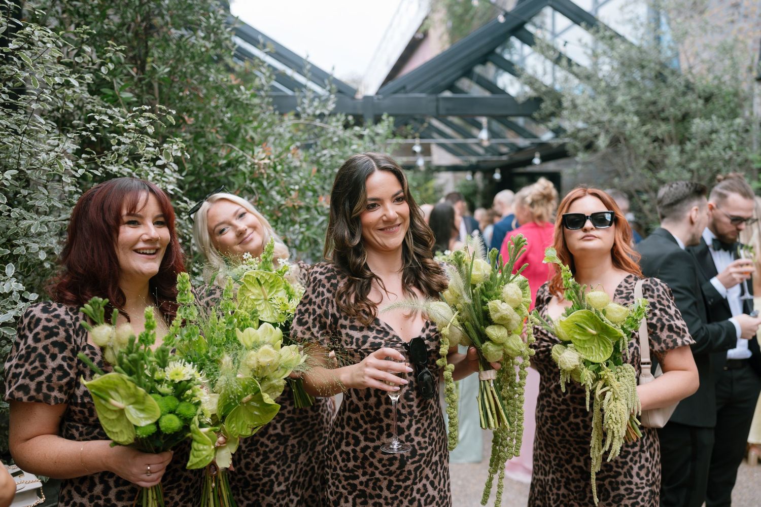 Wedding party members hold large leafy bouquets while celebrating outdoors.