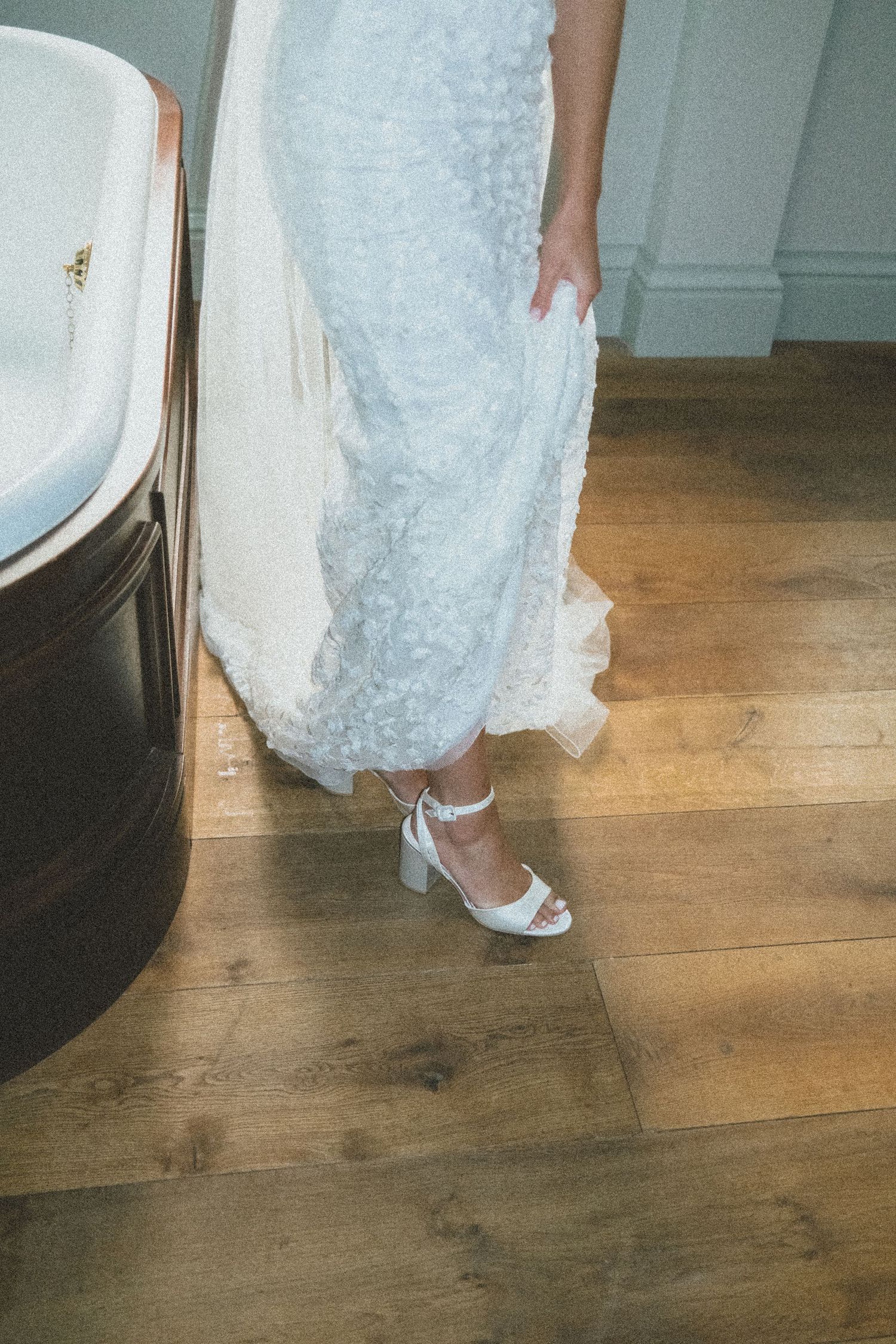 A white lace wedding dress hem and silver strappy heels are shown against a wooden floor.