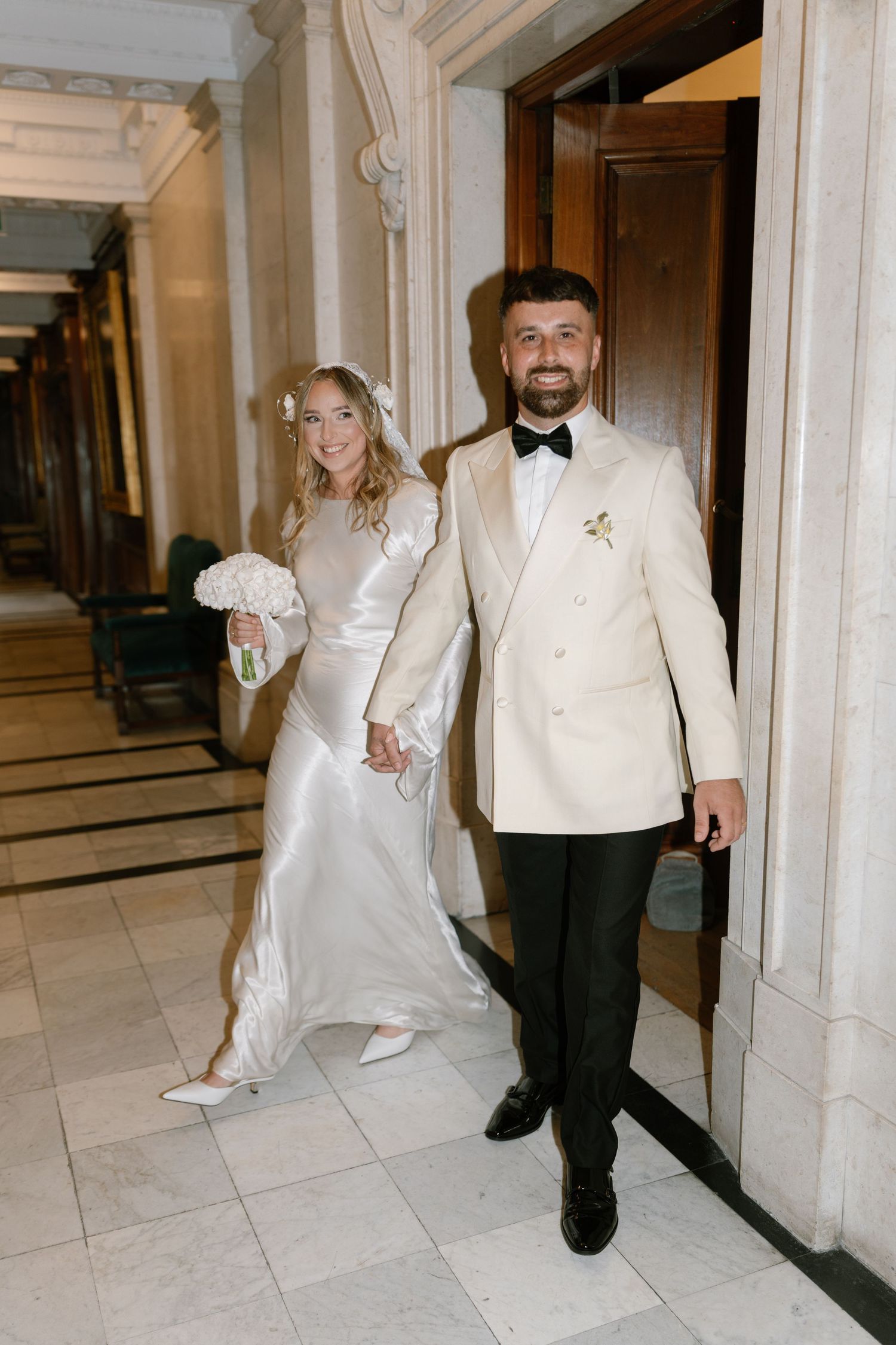 Wedding couple walking through an elegant marble hallway with white columns and tiled floors.