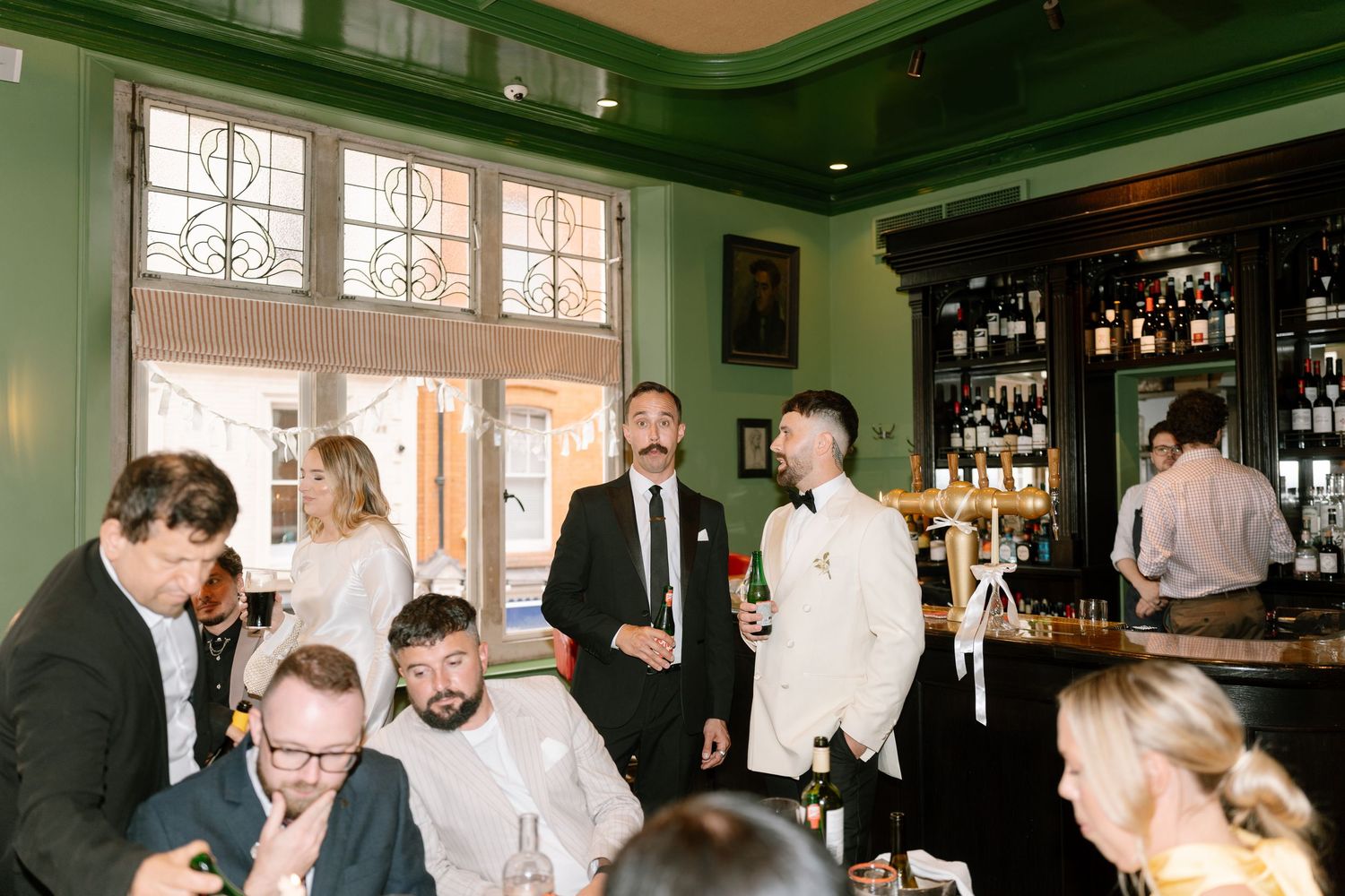 Wedding reception guests mingling in a dark green-walled bar with large windows.